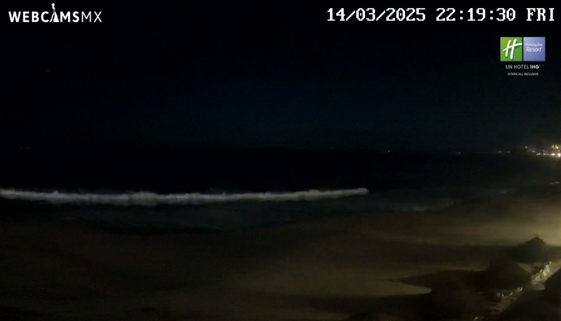 A dark beach at night shows a line of small waves breaking on the shore under a clear, dark sky.