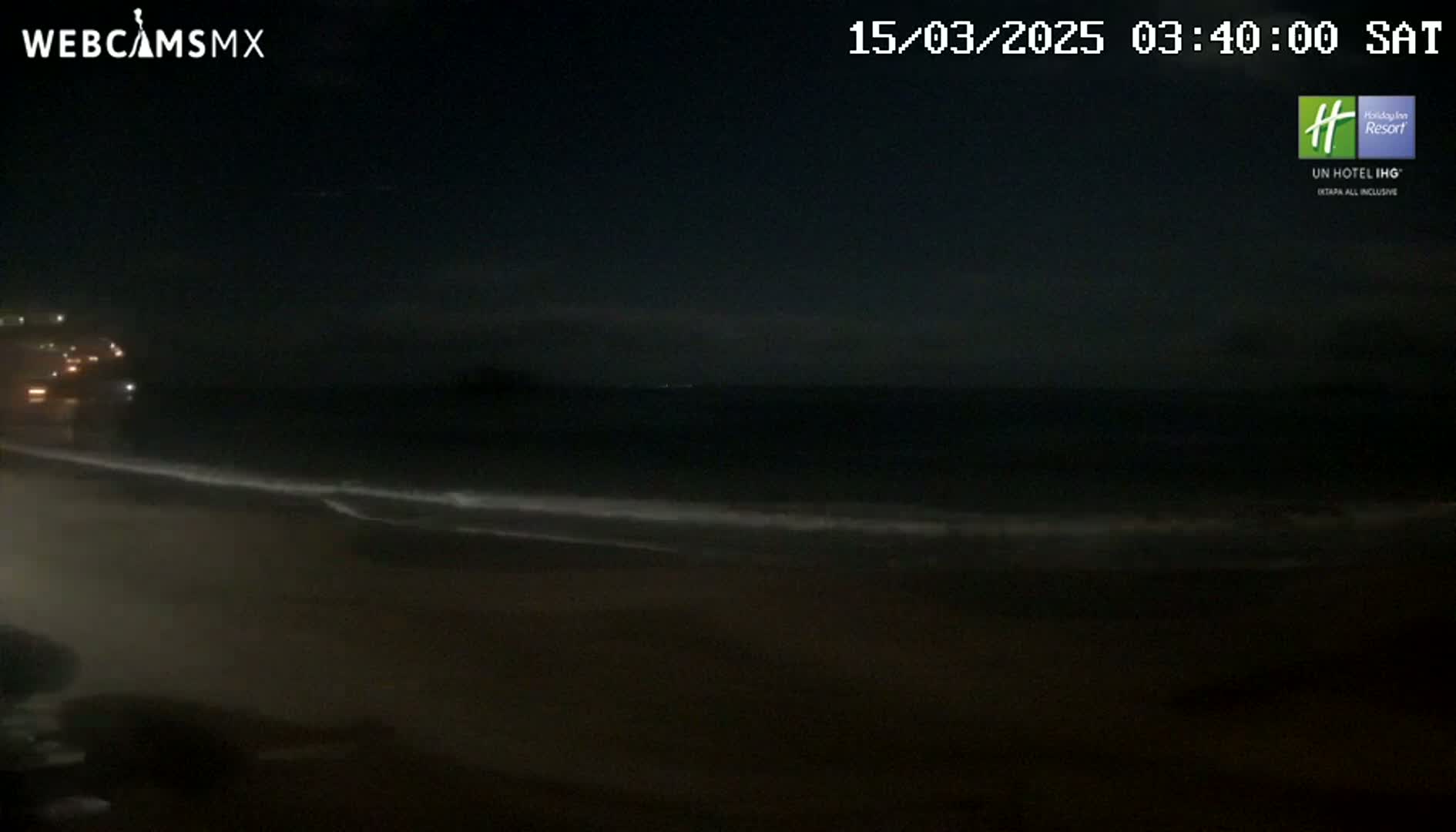 A dark night scene shows a beach with gentle waves lapping the shore under a mostly clear sky.