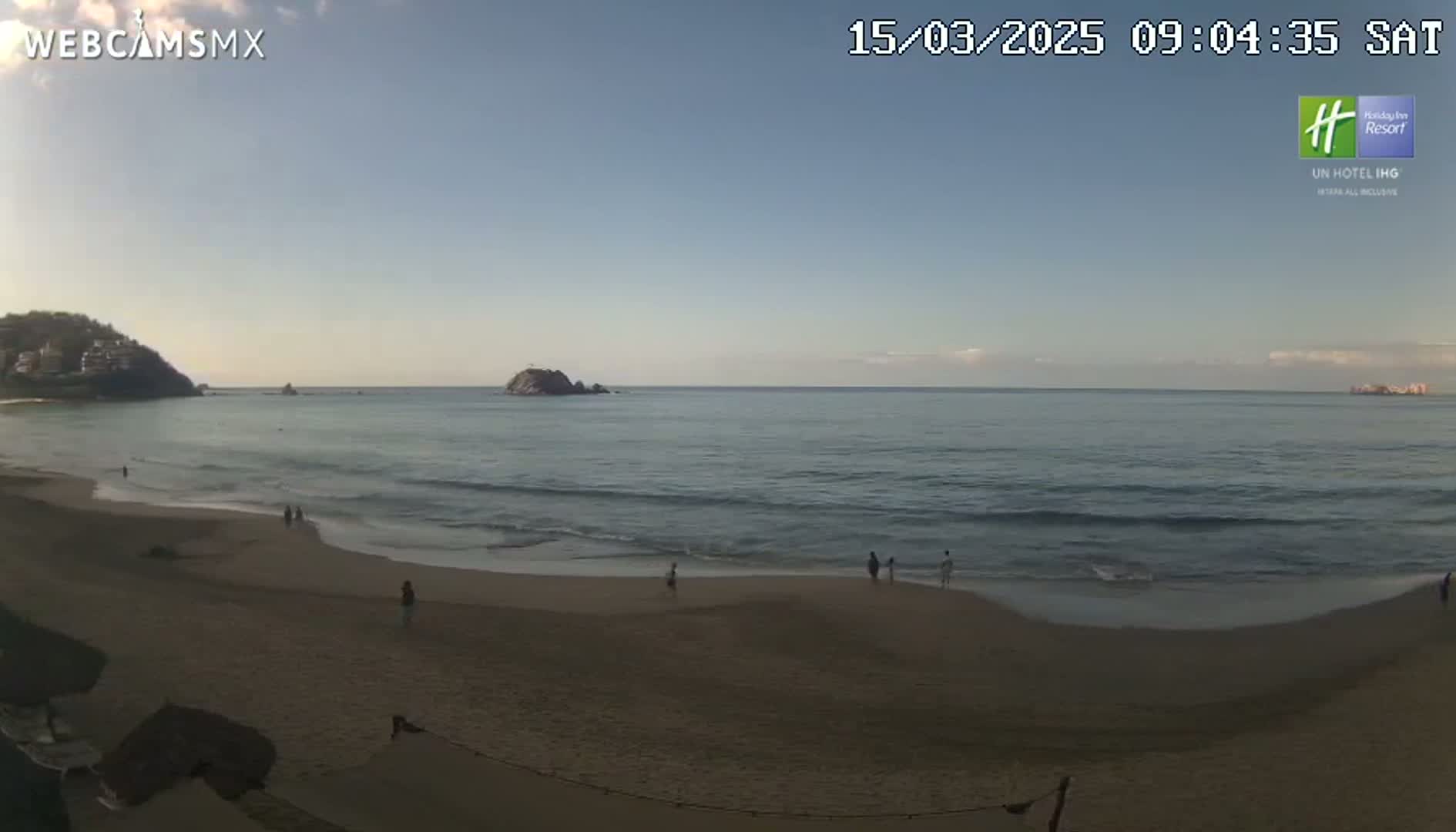 A sandy beach with a few people on it stretches out to a calm ocean with small waves under a clear, bright sky.