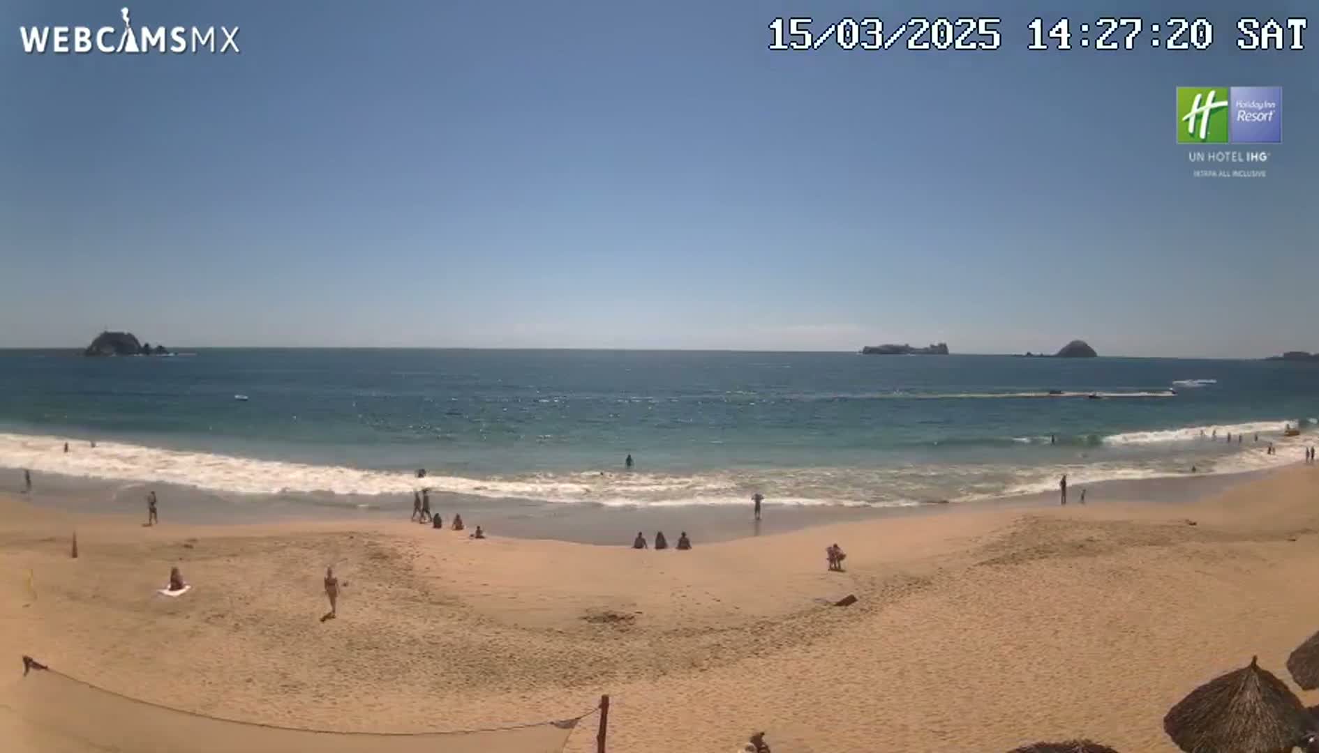 A sunny day at a beach shows calm ocean waves lapping a sandy shore where a few people are swimming and sunbathing.