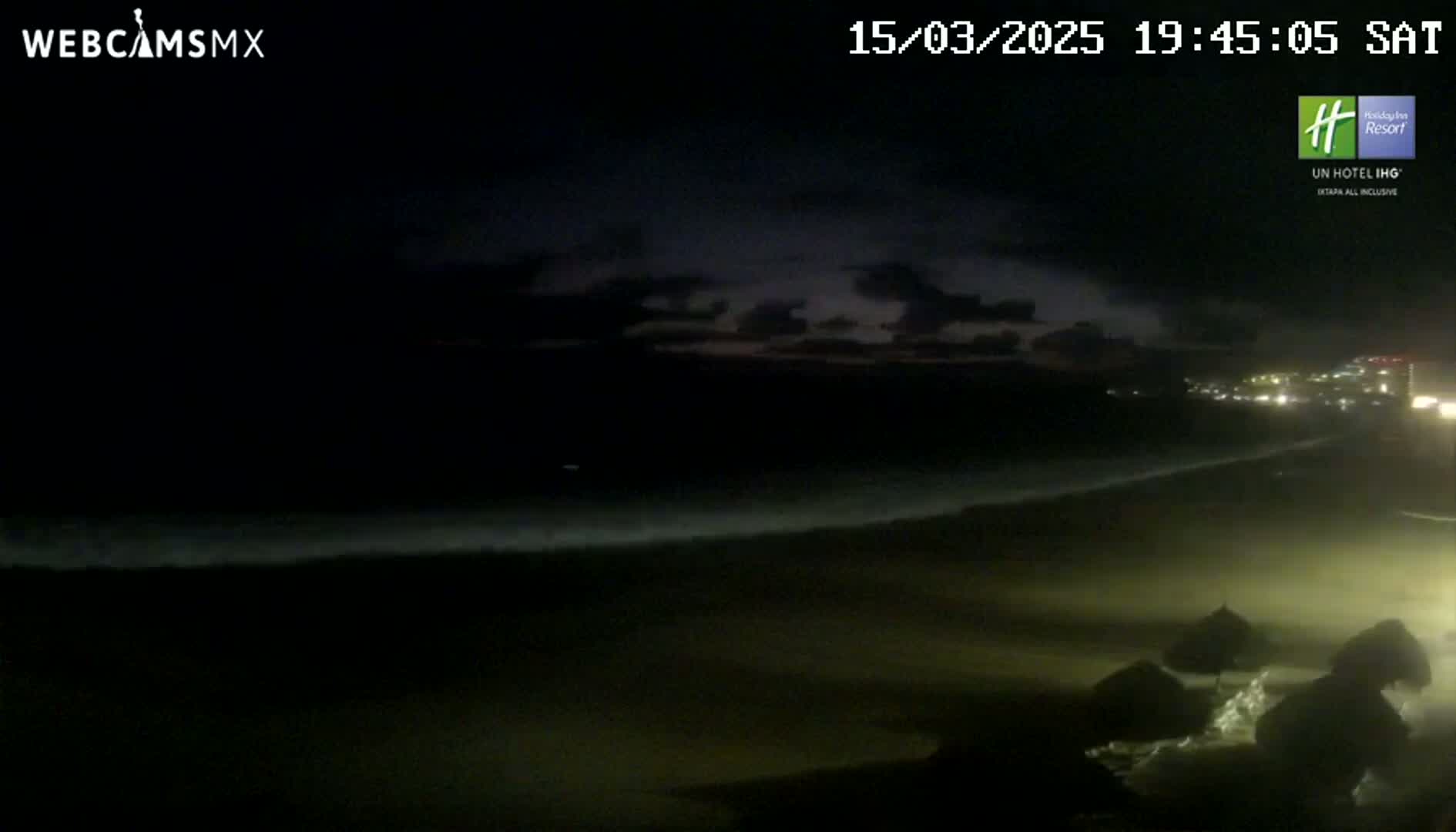 A dark, nighttime beach scene shows waves lapping the shore under a partly cloudy sky.