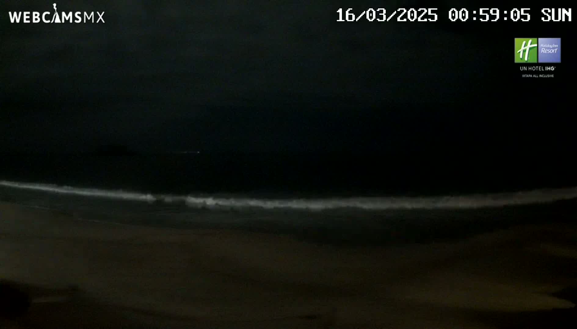 A dark, nighttime beach scene shows gentle waves lapping the shore under a mostly clear sky.