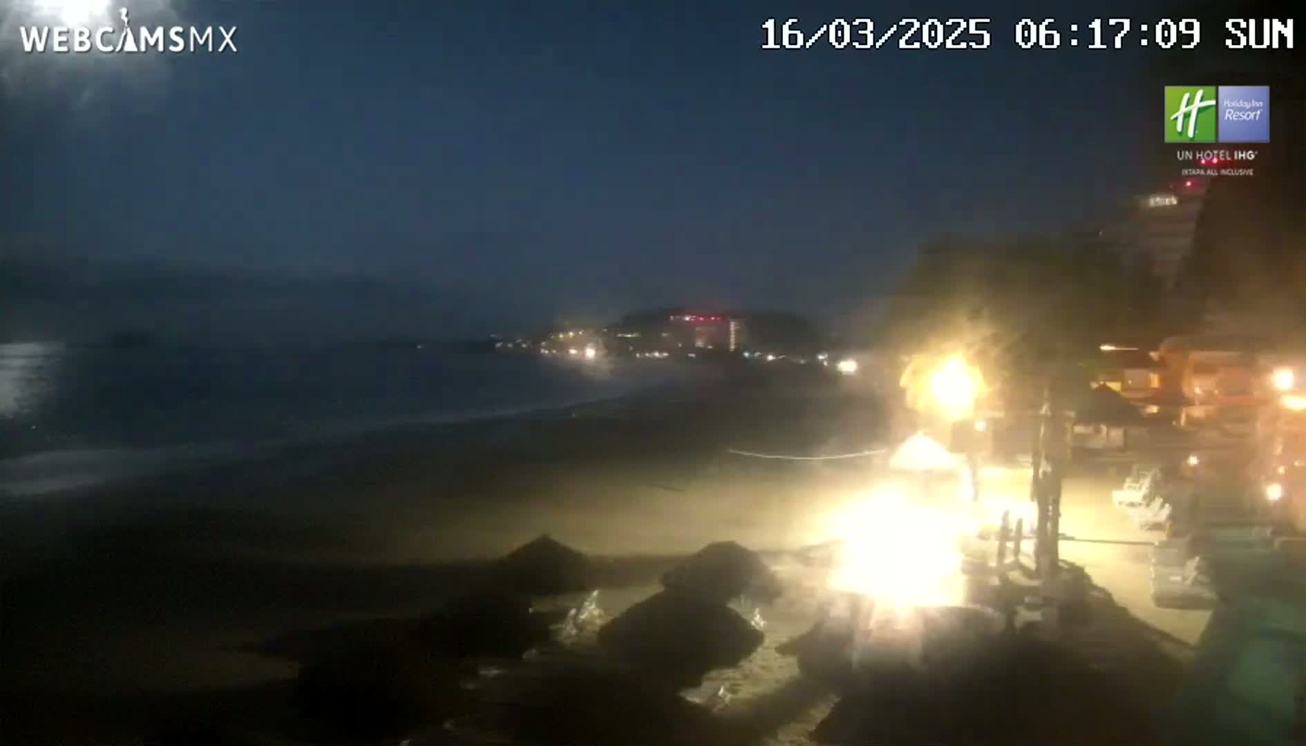 A nighttime beach scene shows dark water lapping at a sandy shore, with lights visible in buildings and on the beach.