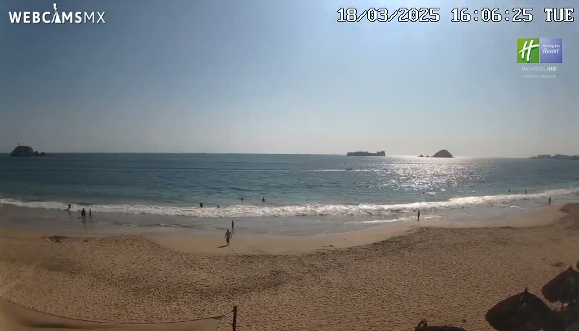 A sandy beach with calm waves and a few people on it stretches towards a calm ocean under a sunny sky, with islands visible in the distance.