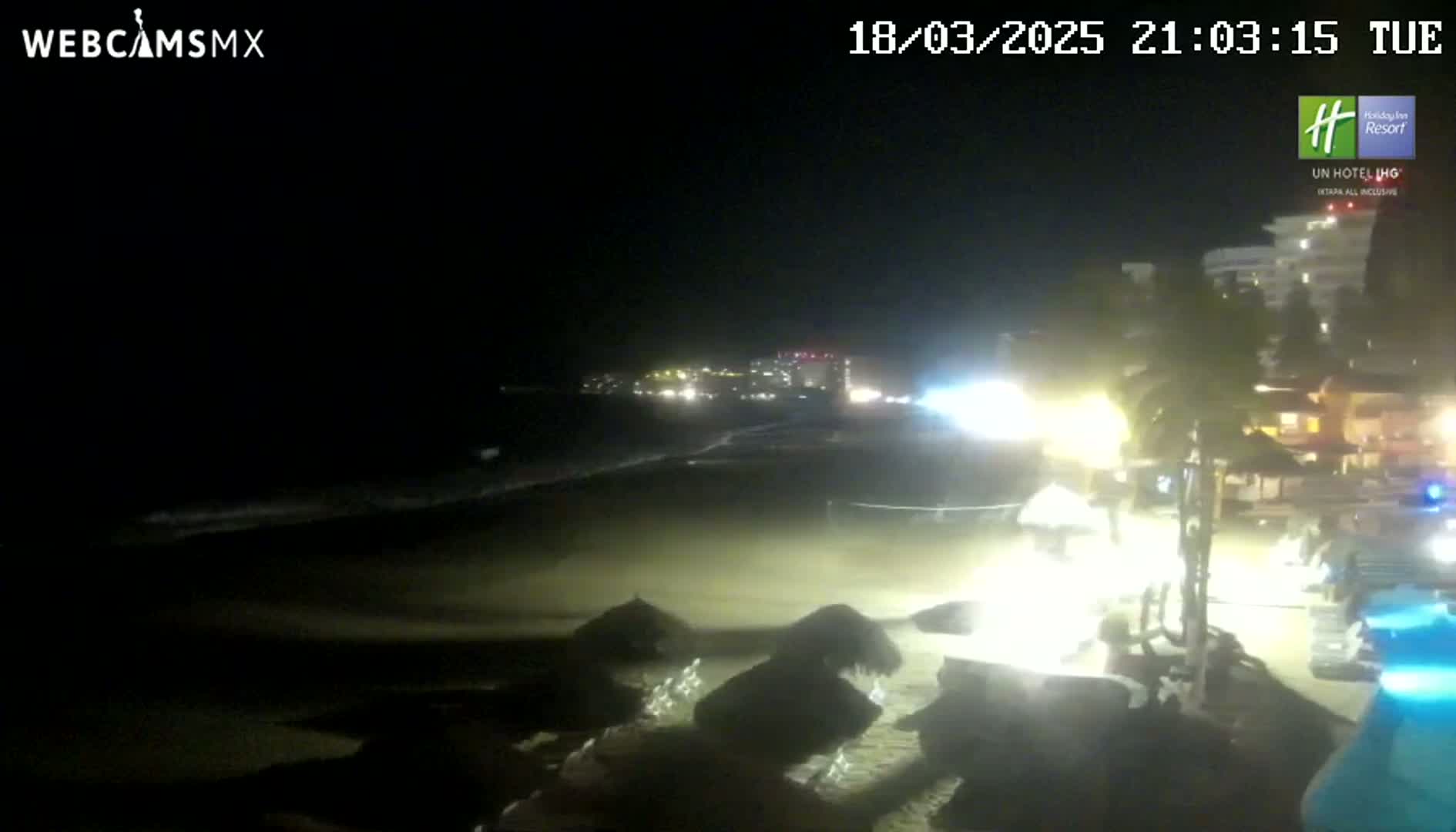 A nighttime beach scene shows gentle waves lapping the shore under a dark sky, with beach umbrellas and some lights visible near buildings in the background.