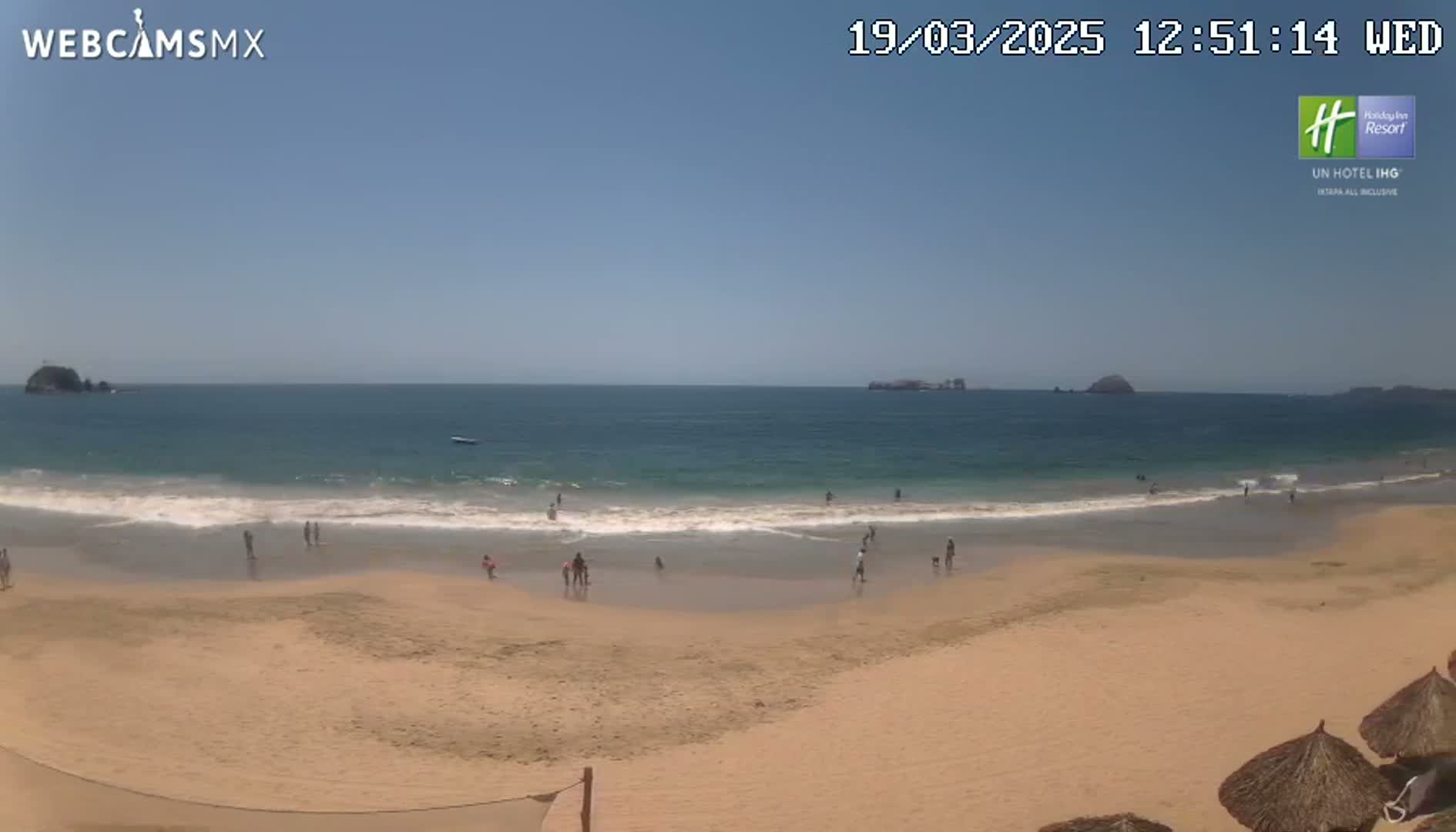 A sandy beach with several people on it, small waves lapping the shore, and islands visible in the distance on a sunny day.