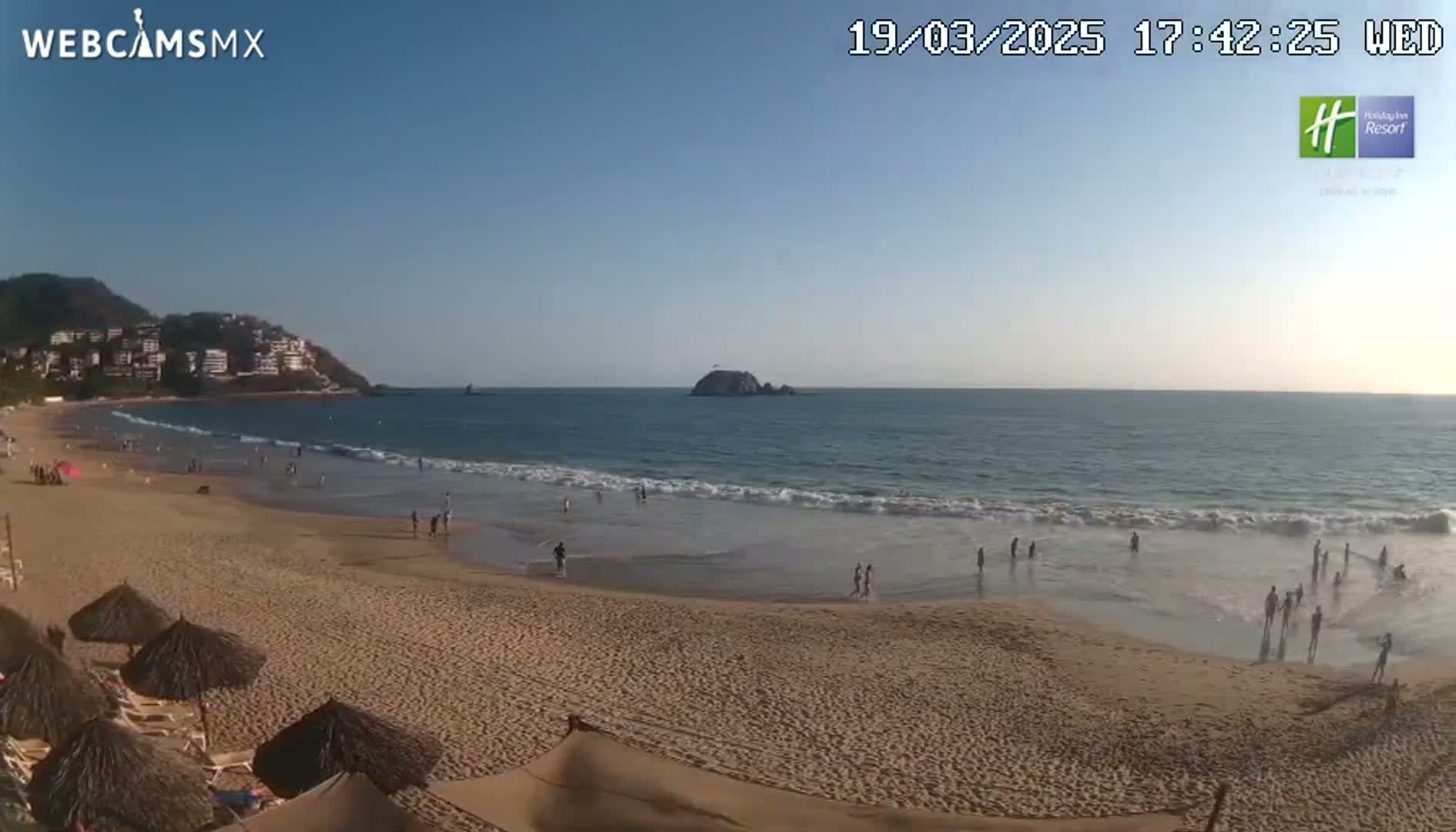 A sandy beach with calm ocean waves under a clear blue sky, showing several people enjoying the water and shore.