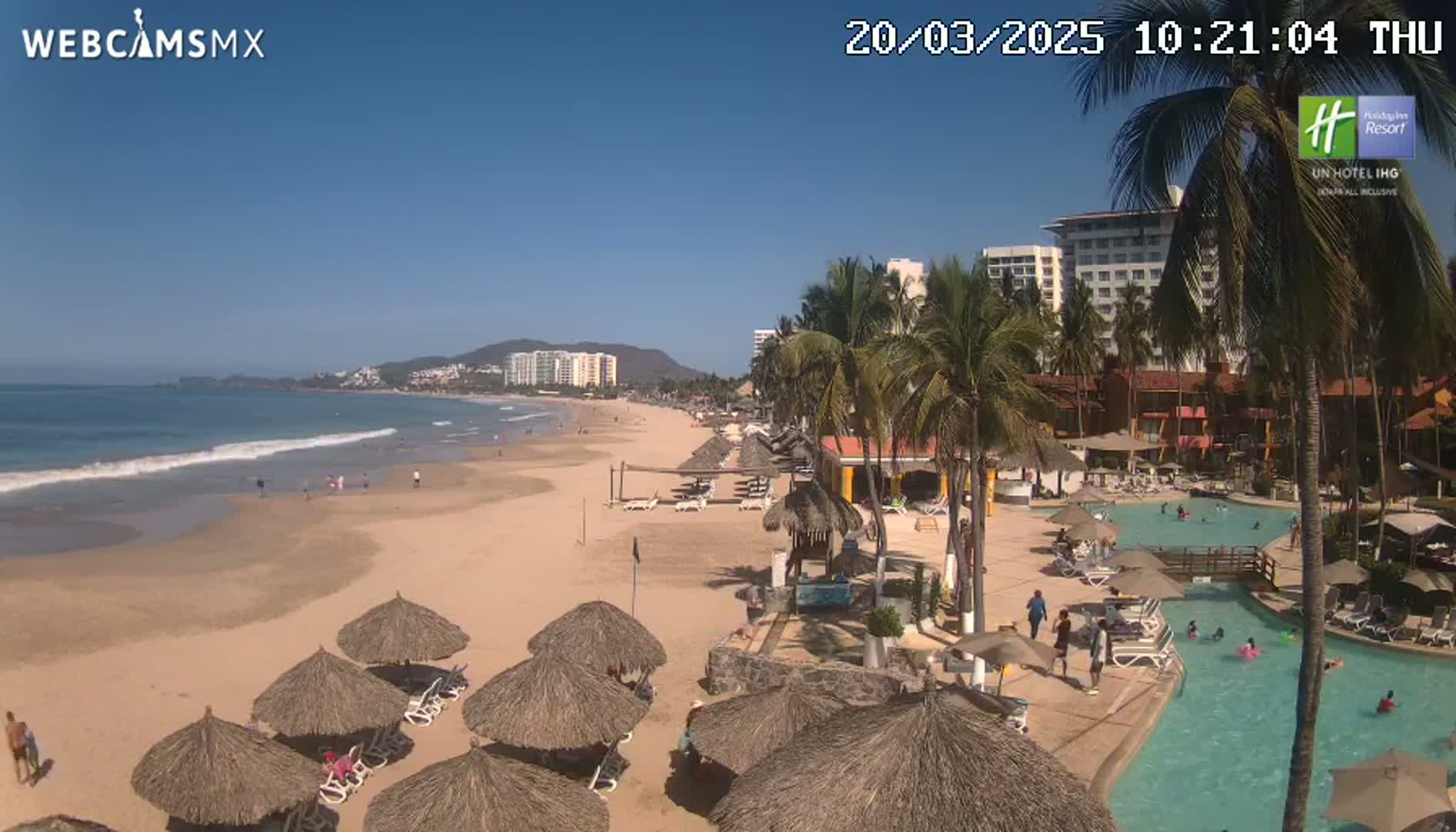 A sandy beach with calm waves, thatched umbrellas, and a resort pool area under a clear blue sky is shown.