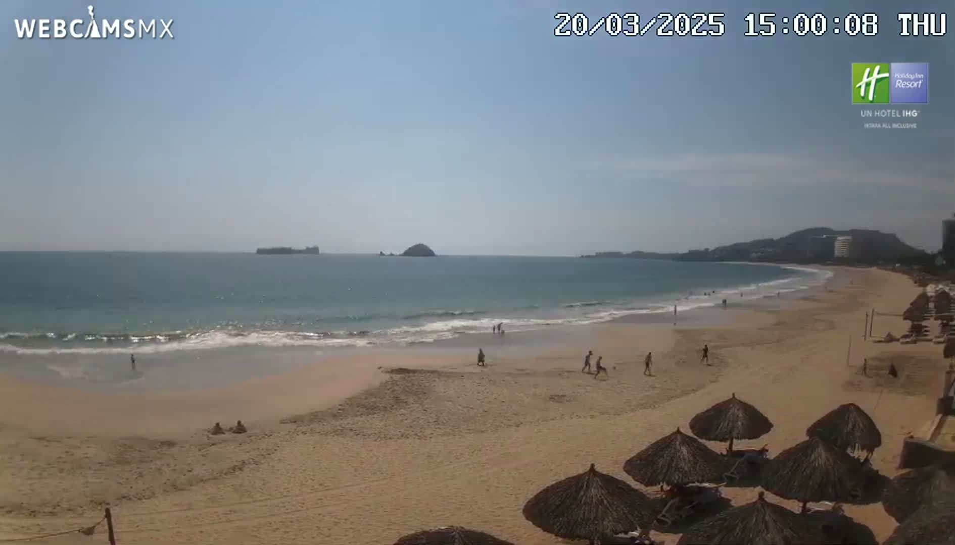 A sandy beach with calm waves, a few people, and several thatched umbrellas under a clear, sunny sky.
