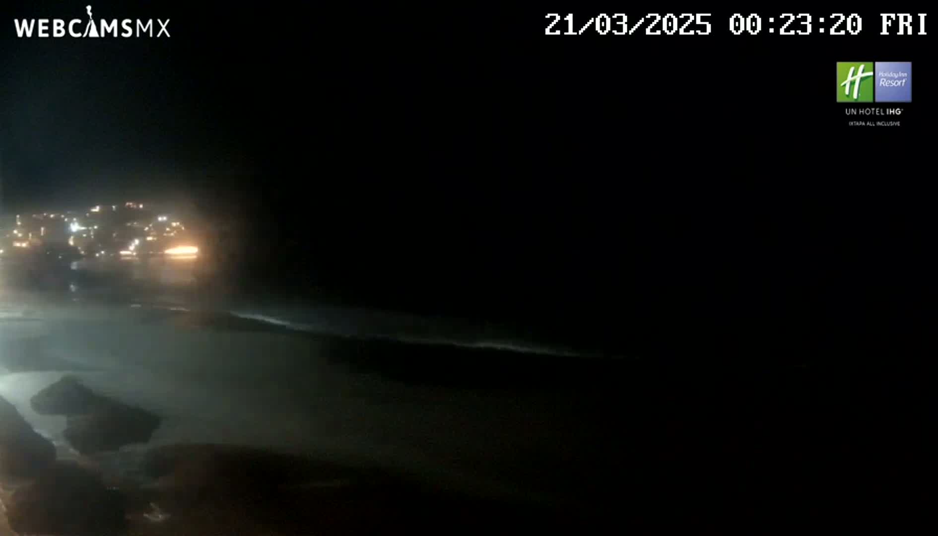 A nighttime coastal scene shows a dark ocean with small waves lapping on the shore, and lights from a town visible in the distance.