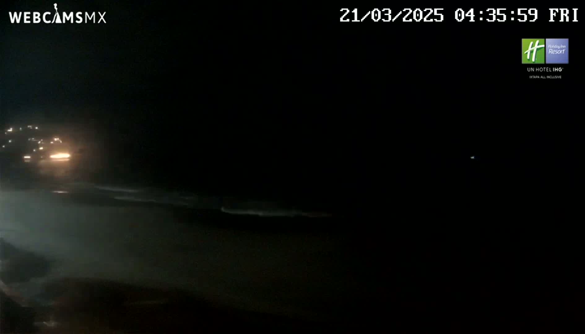 A nighttime view of a dark beach with faint waves and the headlights of a vehicle visible in the distance.
