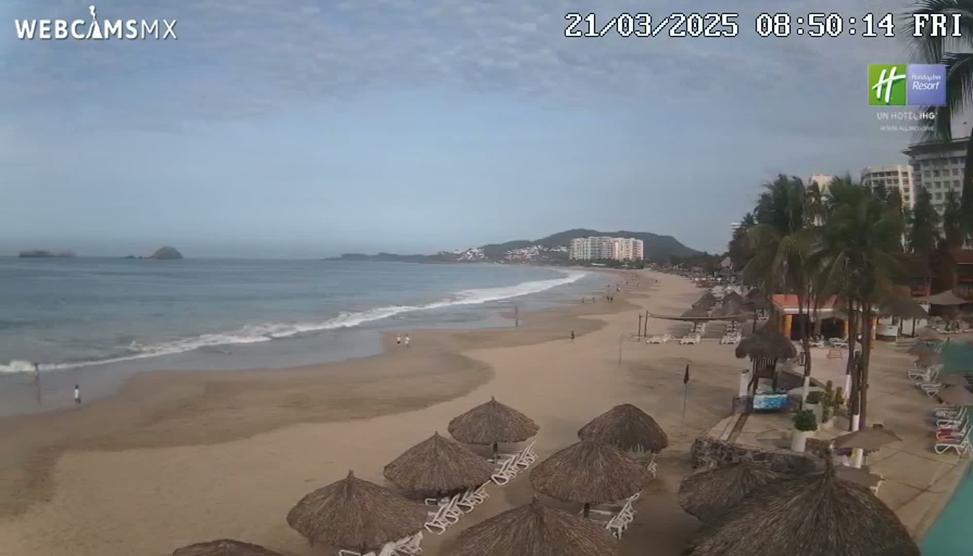 A mostly sunny beach with calm waves, several thatched umbrellas and lounge chairs, and a resort in the background.