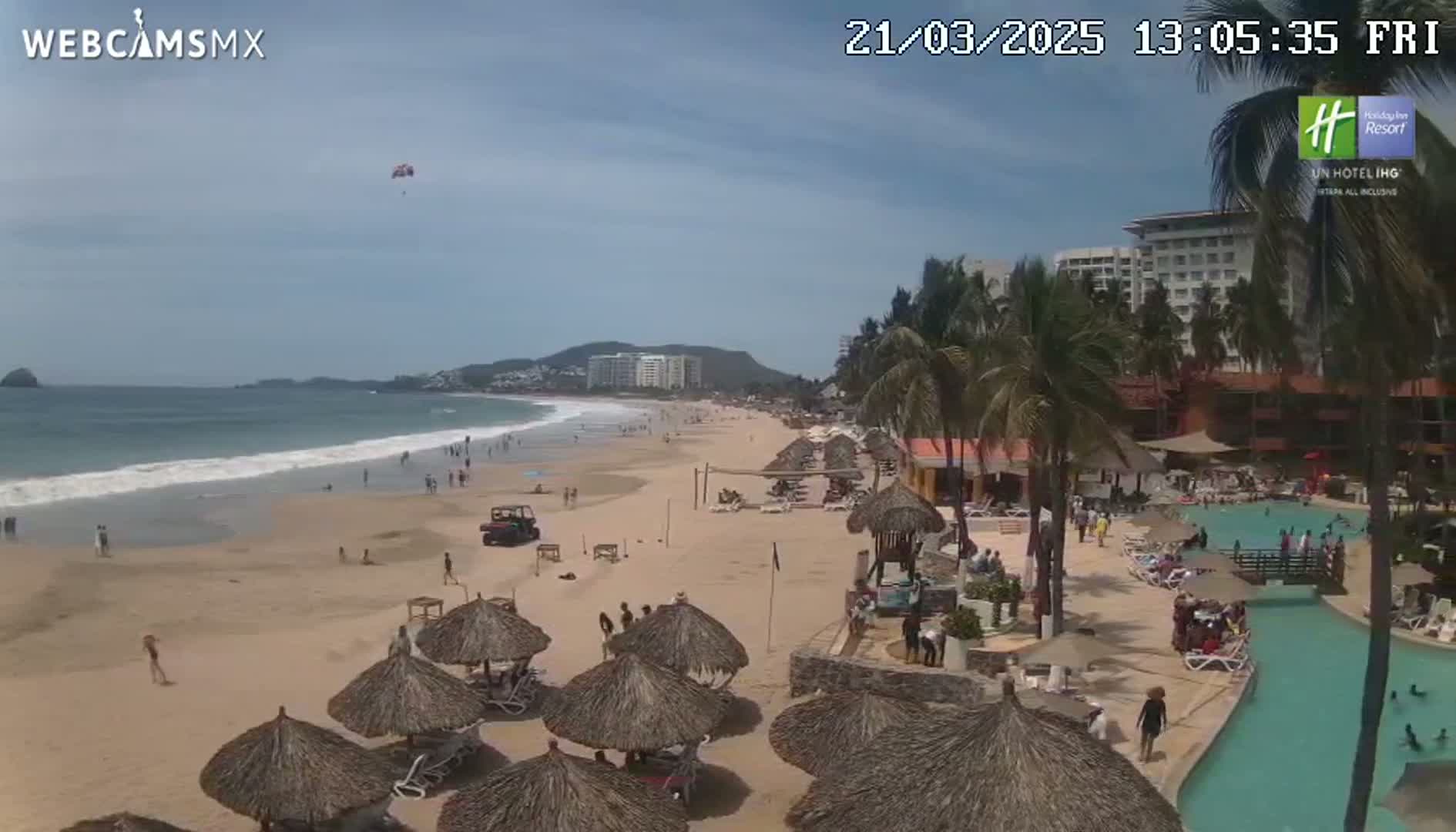A sandy beach with calm waves, thatched umbrellas, a resort, and a parasailor in the sky under sunny conditions.