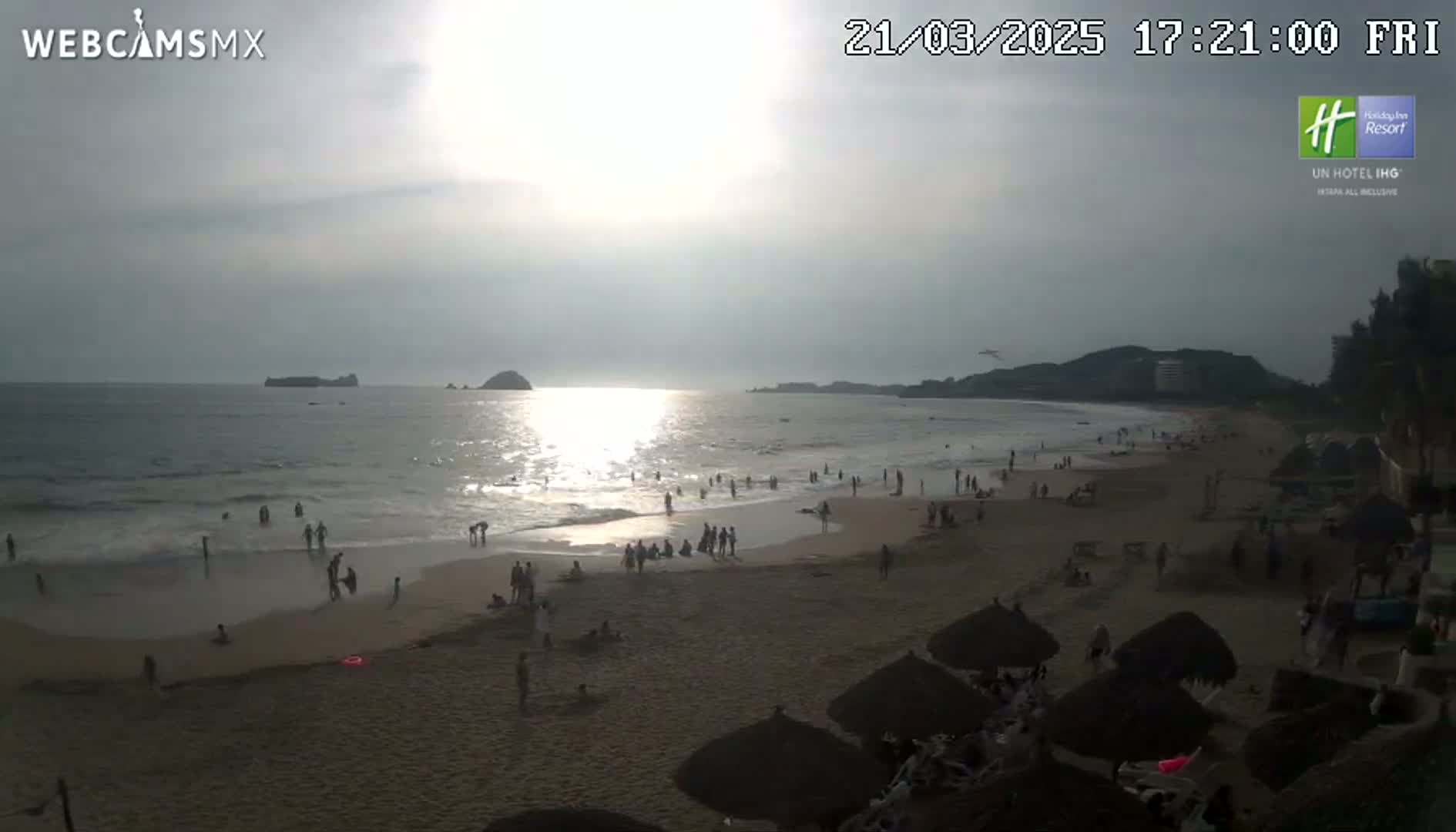 A beach scene at sunset shows many people on the sand and in the water, with a calm ocean and distant islands.