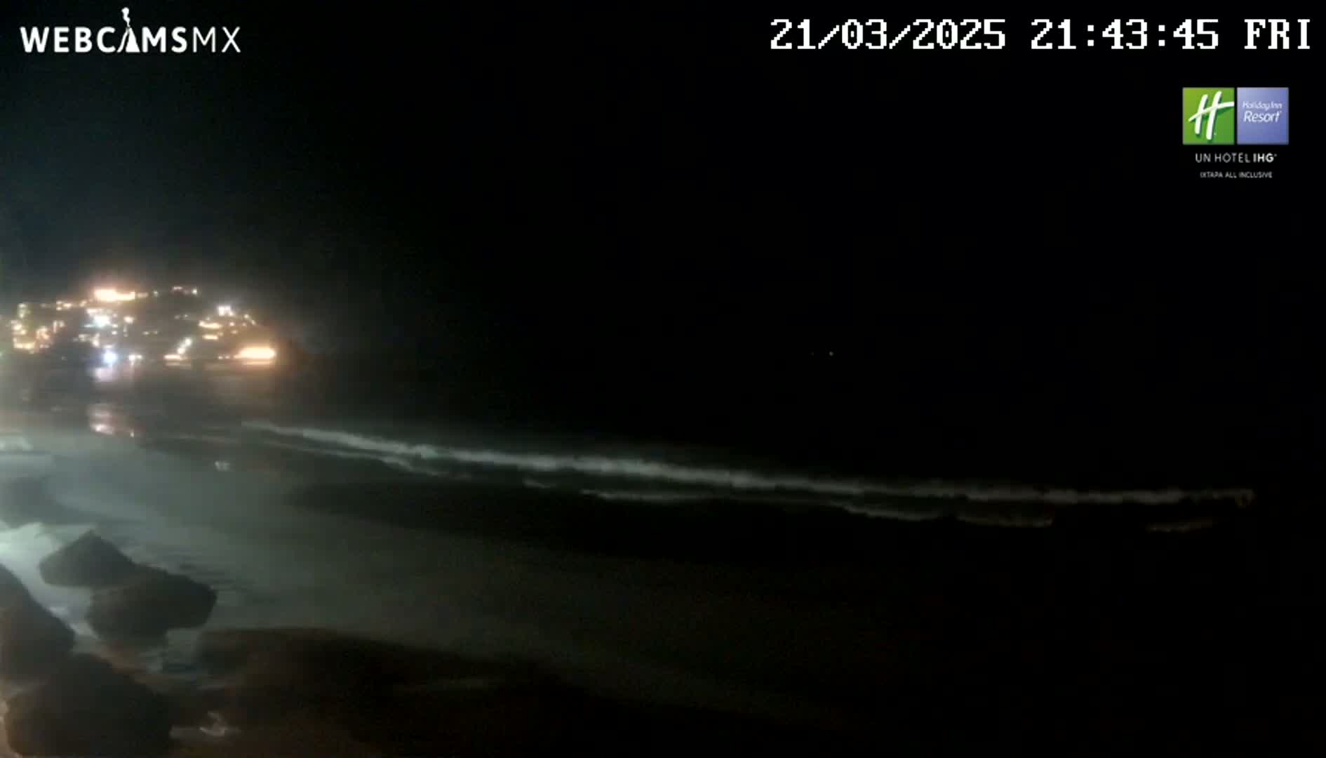 A nighttime beach scene shows dark ocean waves gently lapping the shore near a dimly lit town.