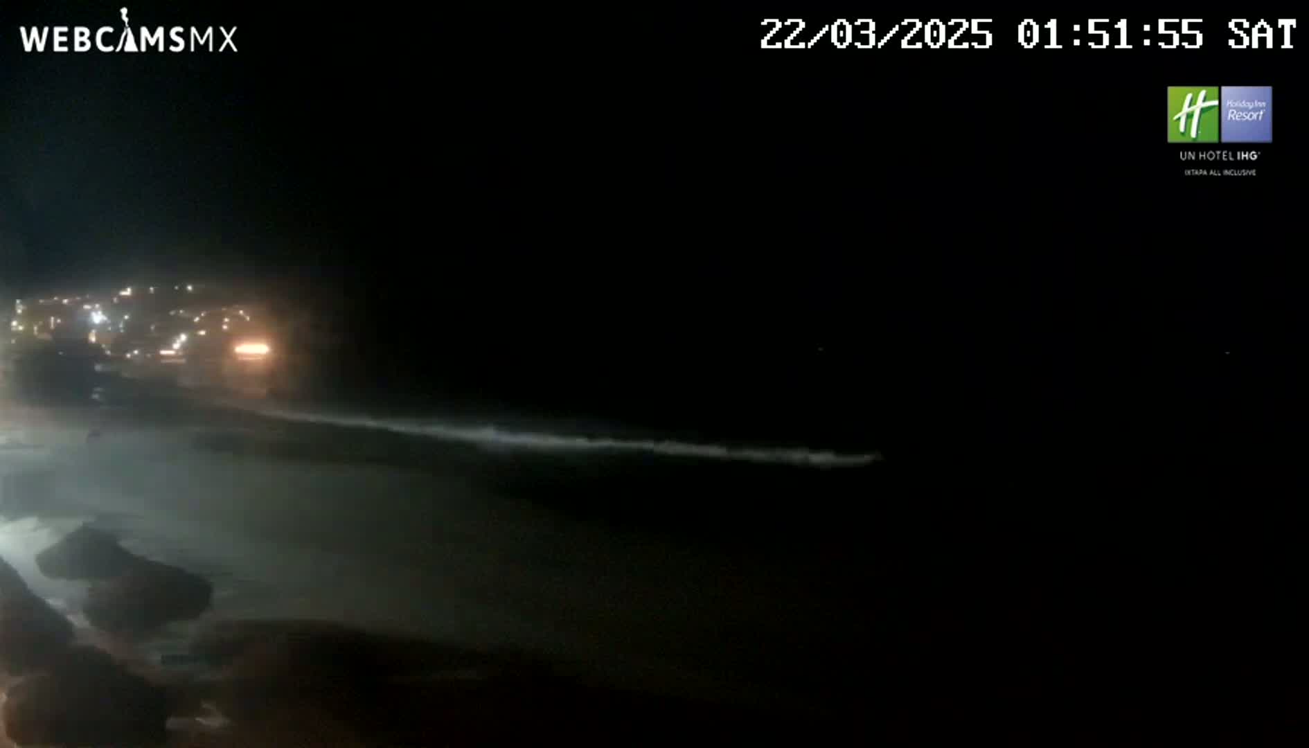 A nighttime view of a beach with gentle waves lapping the shore, illuminated by city lights in the distance.