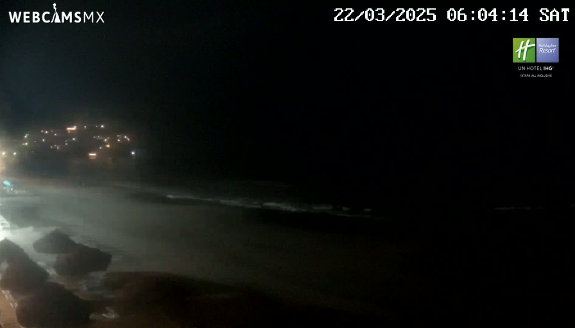 A nighttime beach scene shows dark waves lapping the shore near a dimly lit building in the distance.