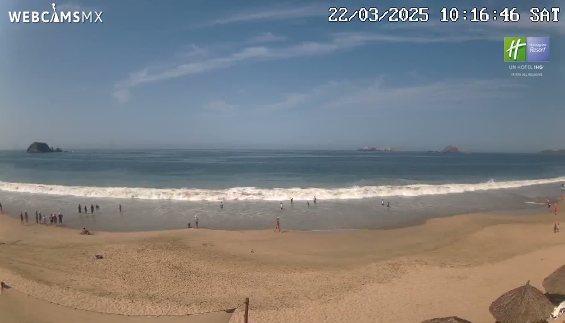 A sandy beach with a few people on it is shown, with small waves breaking on the shore under a clear blue sky.