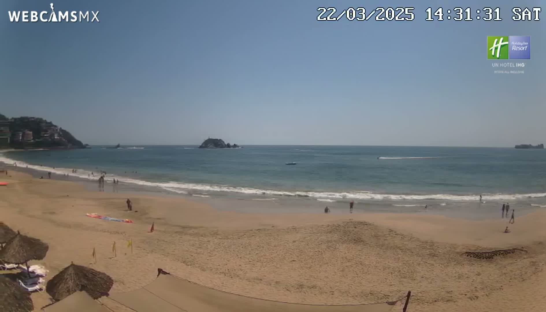 A sandy beach with calm ocean waves, a few people, and thatch umbrellas under a clear blue sky.