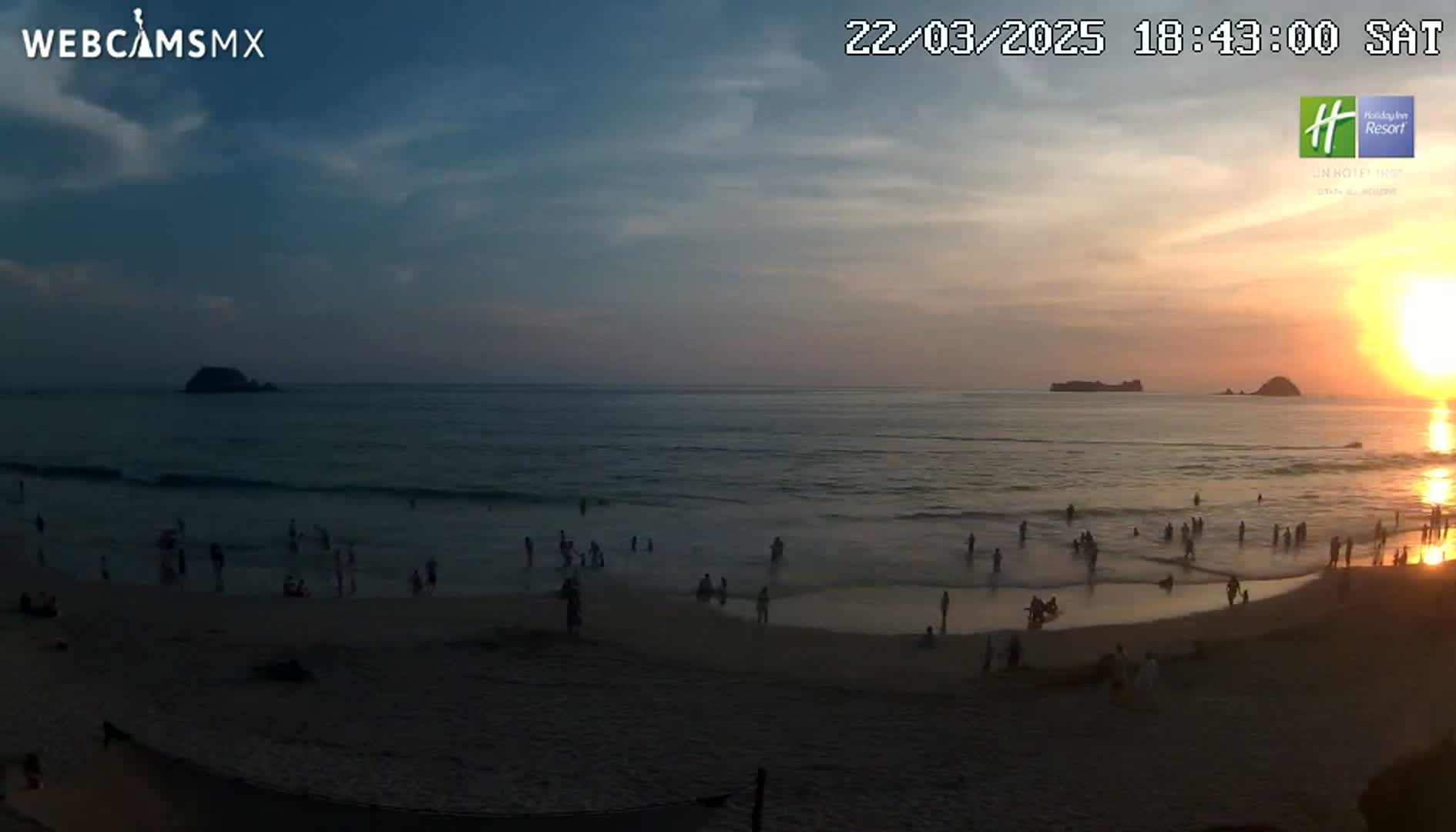 A beach scene at sunset shows people on the sand and in the surf, with a calm ocean and distant islands under a partly cloudy sky.