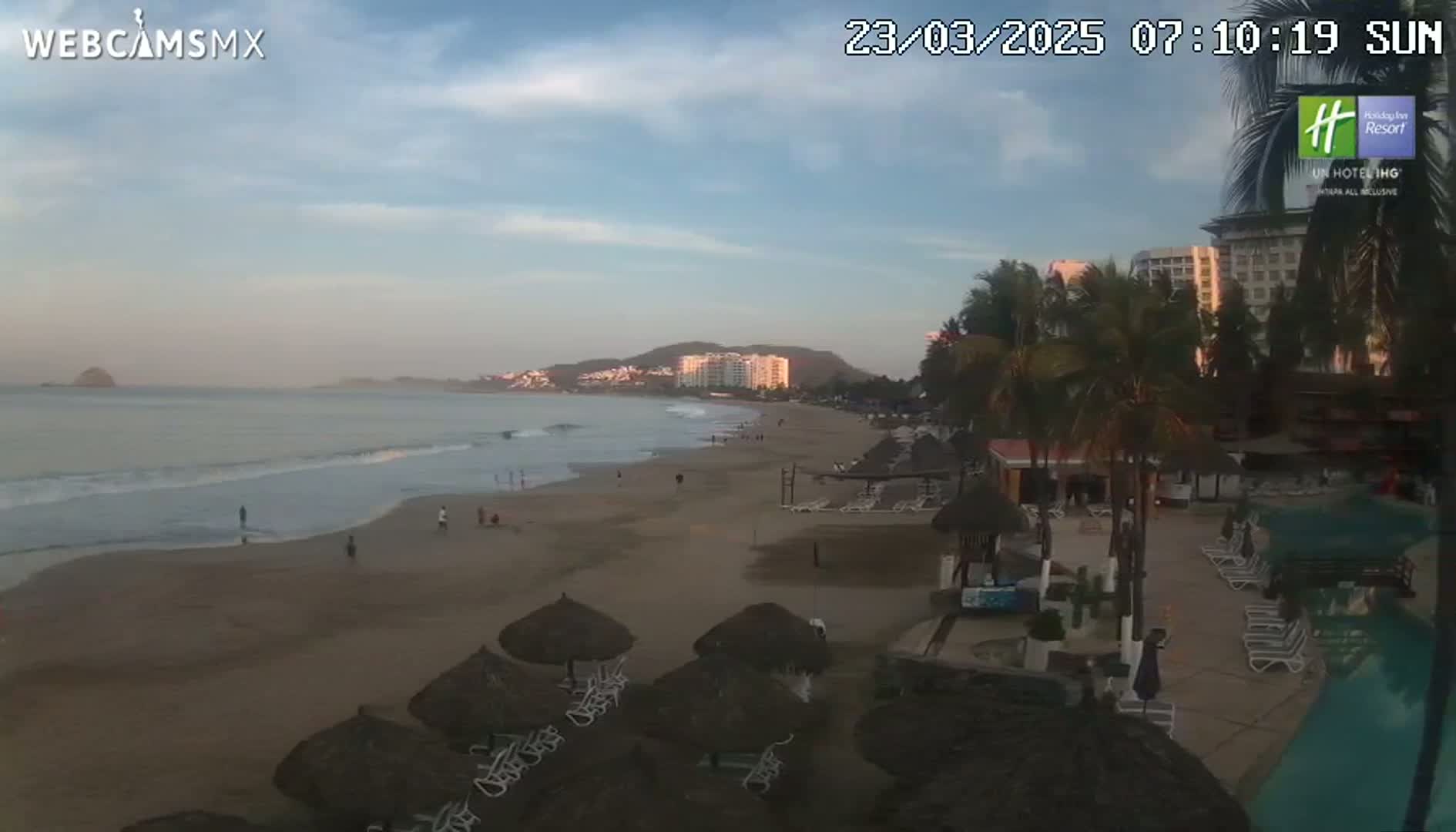 A sandy beach with calm waves and a few people is seen next to a resort with palm trees under a partly cloudy sky.