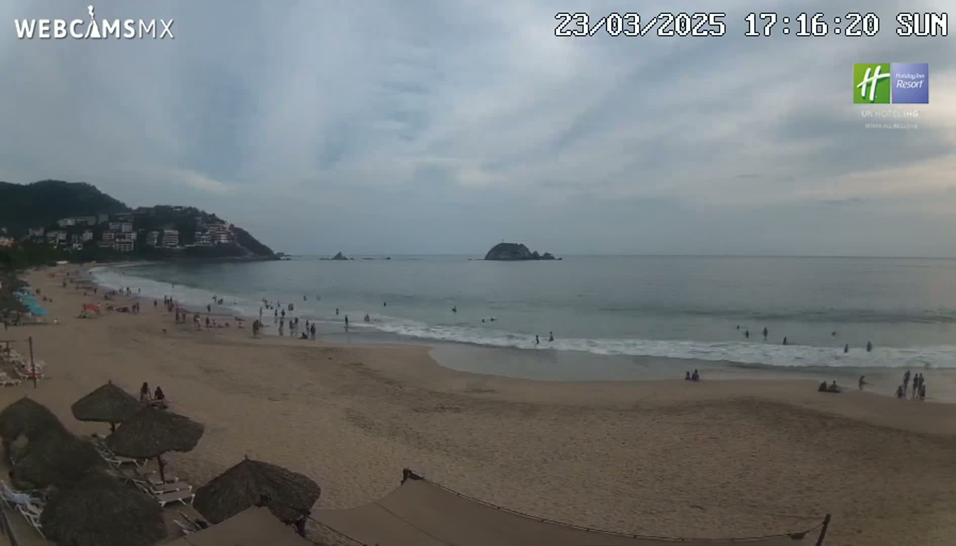 A sandy beach with scattered people and umbrellas under an overcast sky, overlooks a calm ocean with a small island in the distance, backed by a hillside with buildings.
