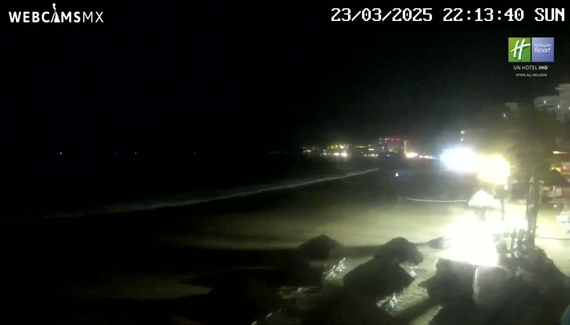 A nighttime beach scene shows dark, gently lapping waves on a sandy shore, with lights illuminating a section of the beach near some rocks and a distant city skyline.