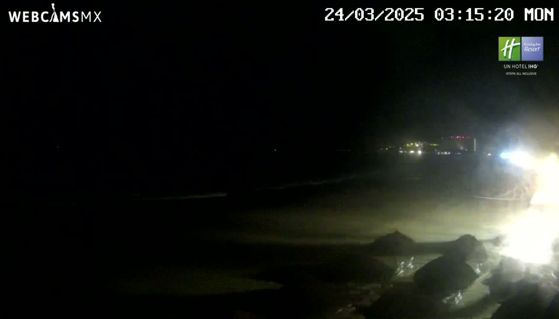 A dark, nighttime beach scene shows a dimly lit shoreline with rocks in the foreground and distant lights visible across the water.