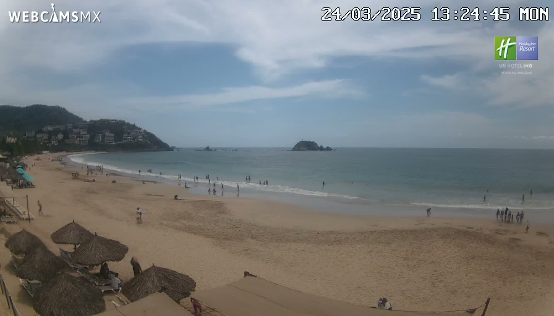A sandy beach with scattered people and beach umbrellas stretches out to a calm ocean under a partly cloudy sky.