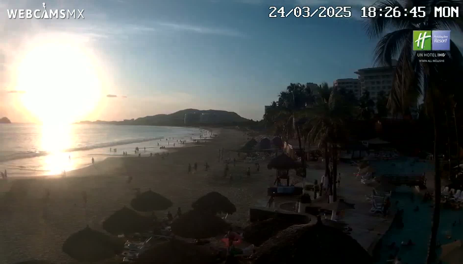 A beach scene at sunset shows a sandy shore with scattered people, palm trees, buildings, and a swimming pool, under a mostly clear sky.