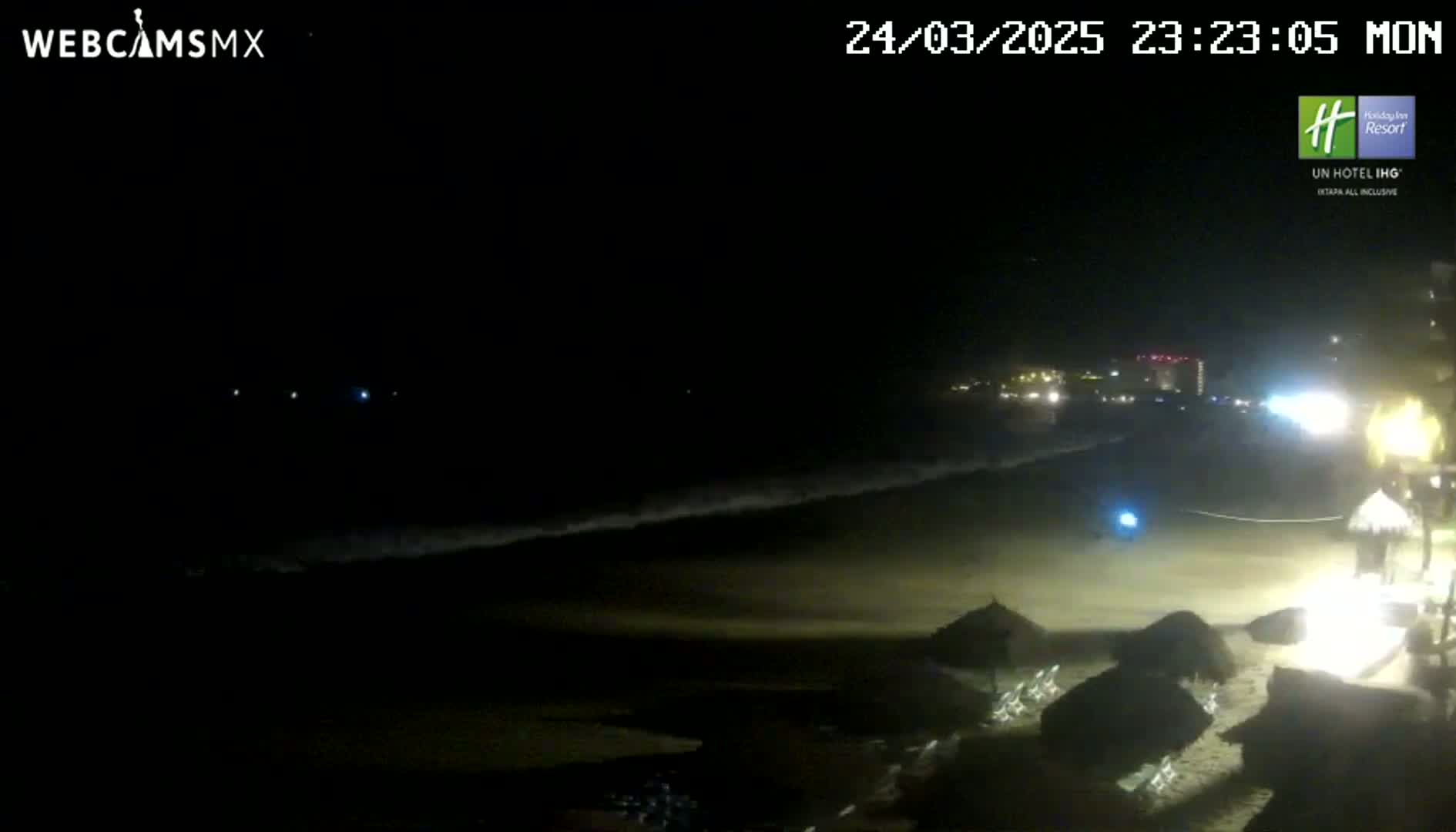 A dark, nighttime beach scene shows a mostly empty sandy shore with several beach umbrellas, gentle waves, and distant lights from buildings along the coast.