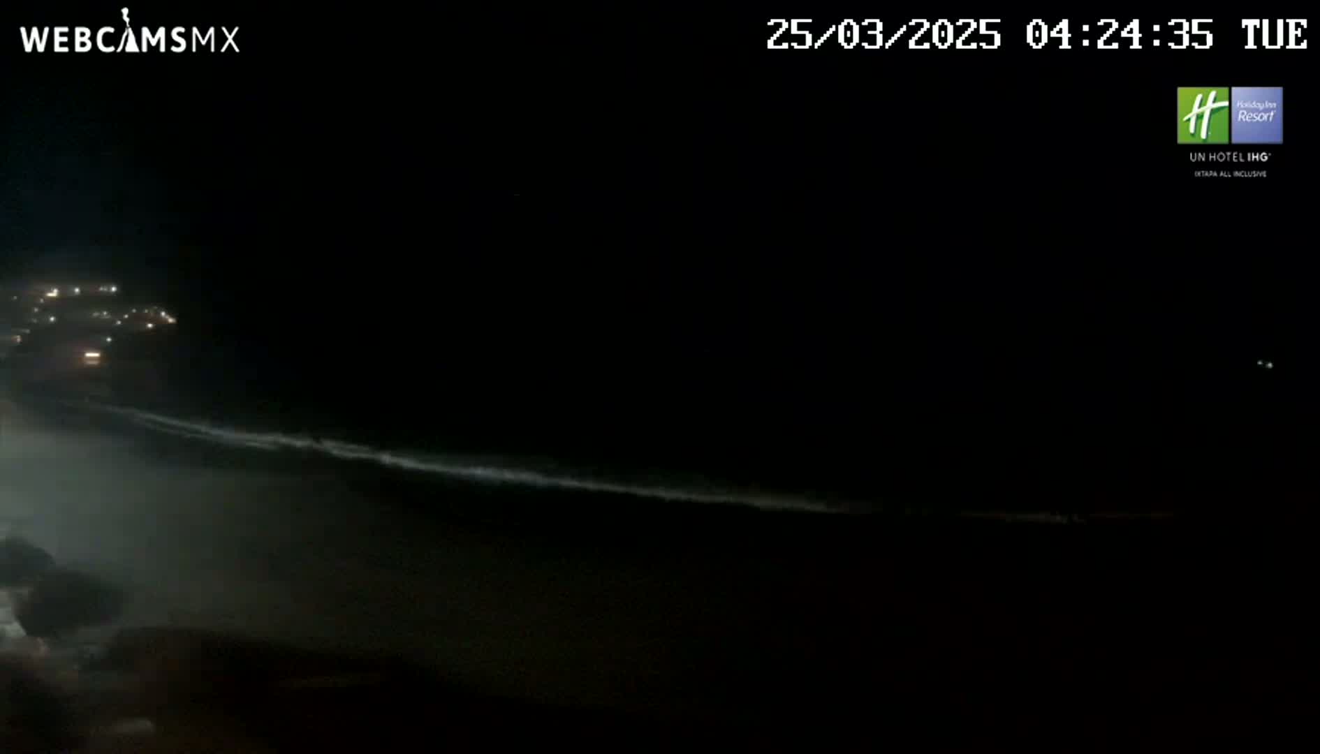 A nighttime beach scene shows a dark ocean with small waves and faintly lit buildings in the distance under a clear, dark sky.