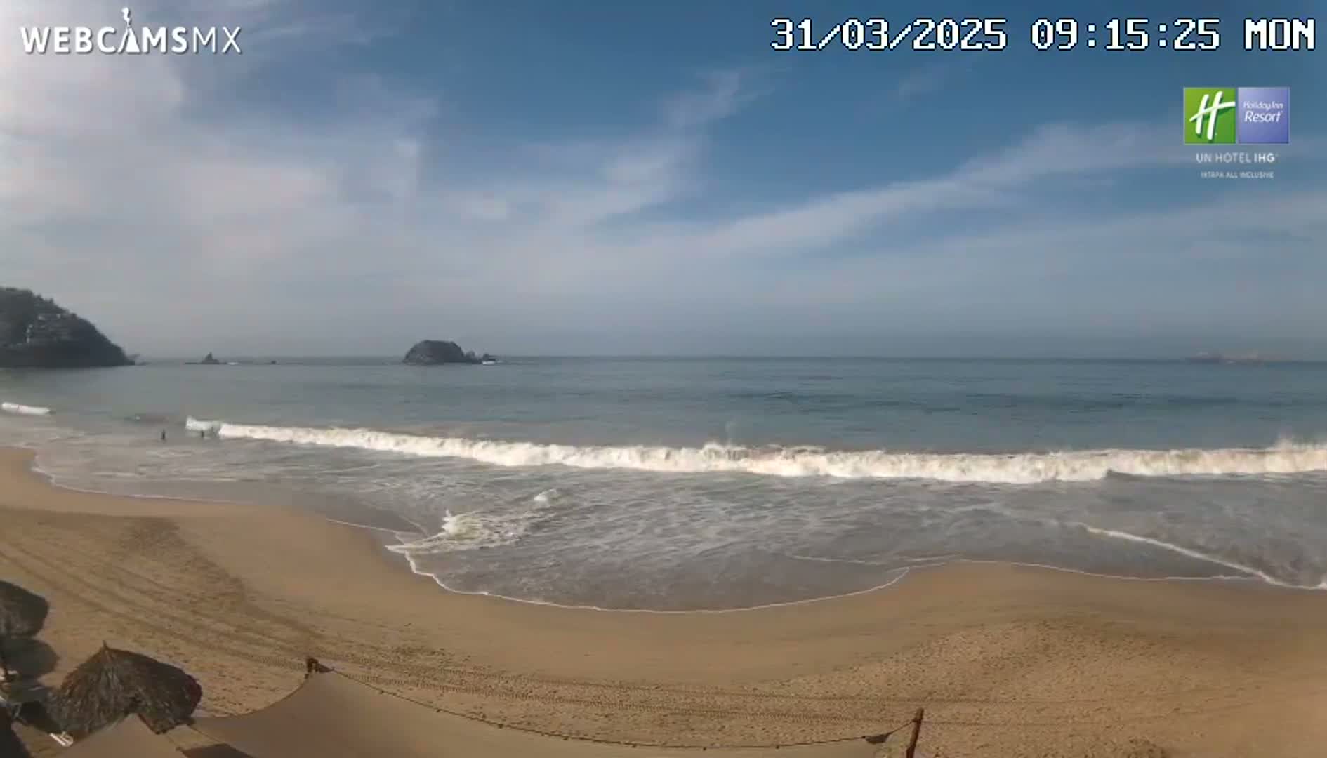 A sandy beach with gentle waves rolls onto the shore under a partly cloudy sky.