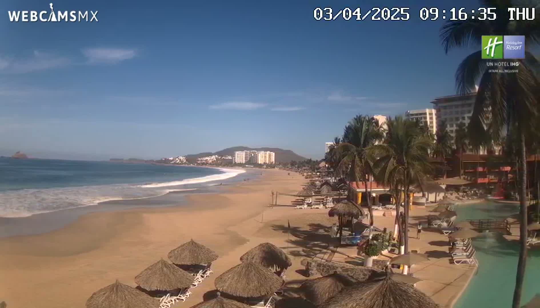 A sandy beach with calm waves stretches toward a line of hotels and palm trees under a clear blue sky.