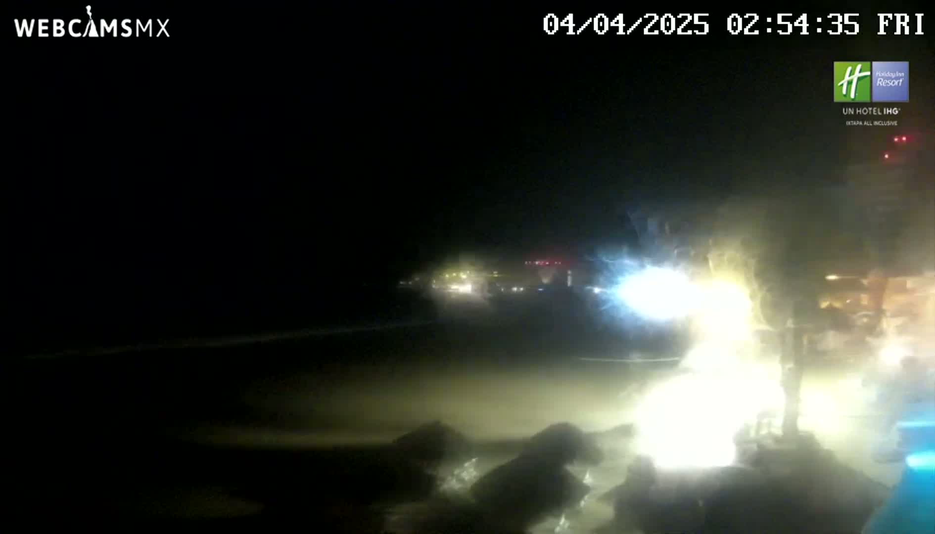 A nighttime beach scene shows a brightly lit area near the water, with blurry lights from buildings and vehicles in the background under a dark sky.
