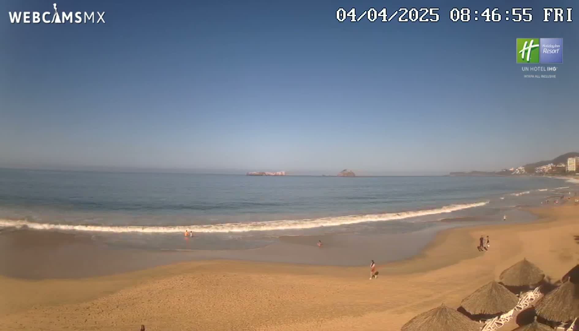 A sandy beach with gentle waves under a clear blue sky, a few people are visible on the beach and in the ocean.