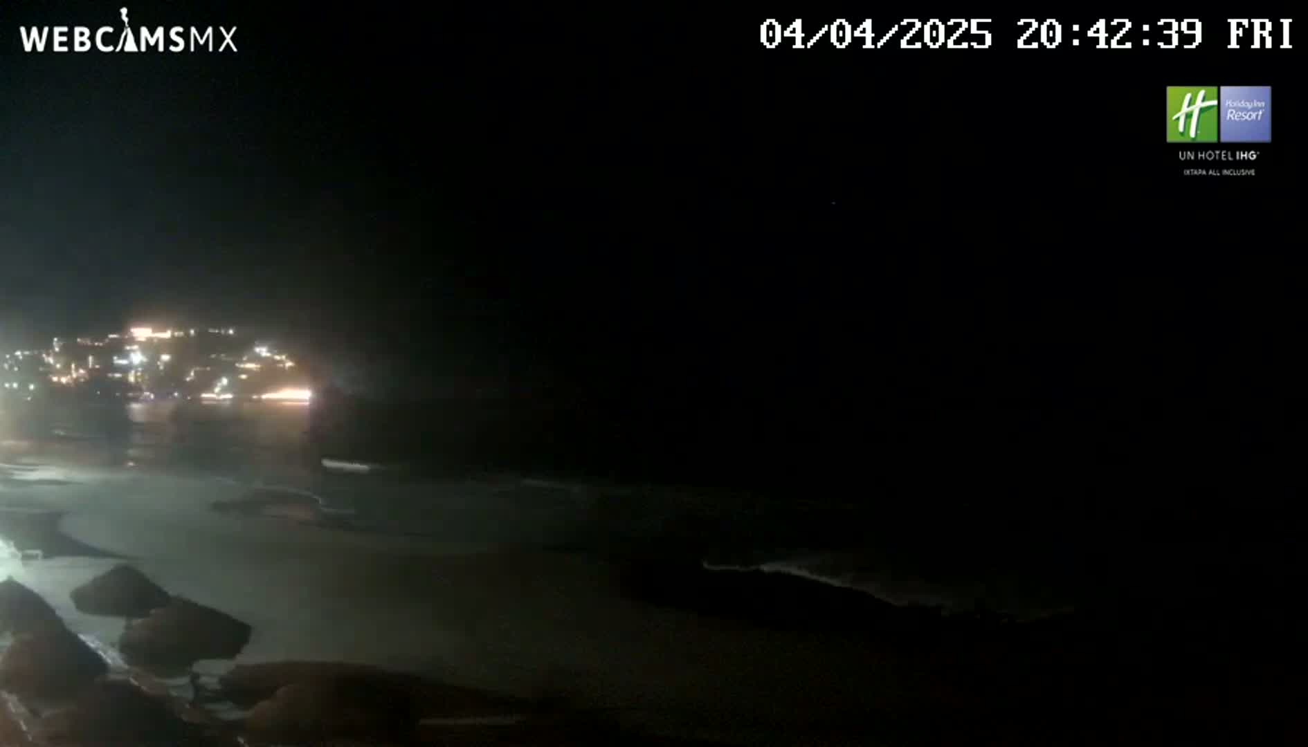 A nighttime view of a dark beach with gently lapping waves and a distant town illuminated by lights.