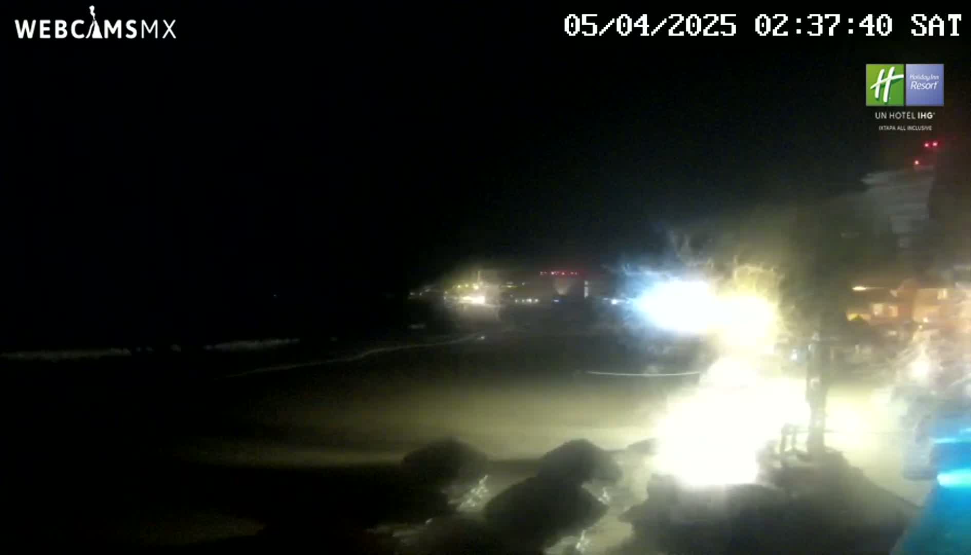 A nighttime beach scene shows softly lapping waves, dimly lit buildings in the distance, and brightly lit vegetation near the camera.