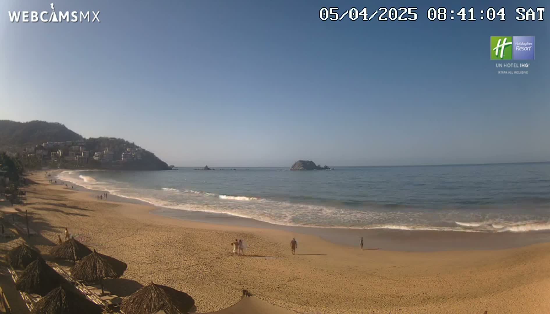 A sandy beach with a few people walking on it, under a clear blue sky,  meets a calm ocean with gentle waves lapping the shore, with hills and buildings visible in the background.