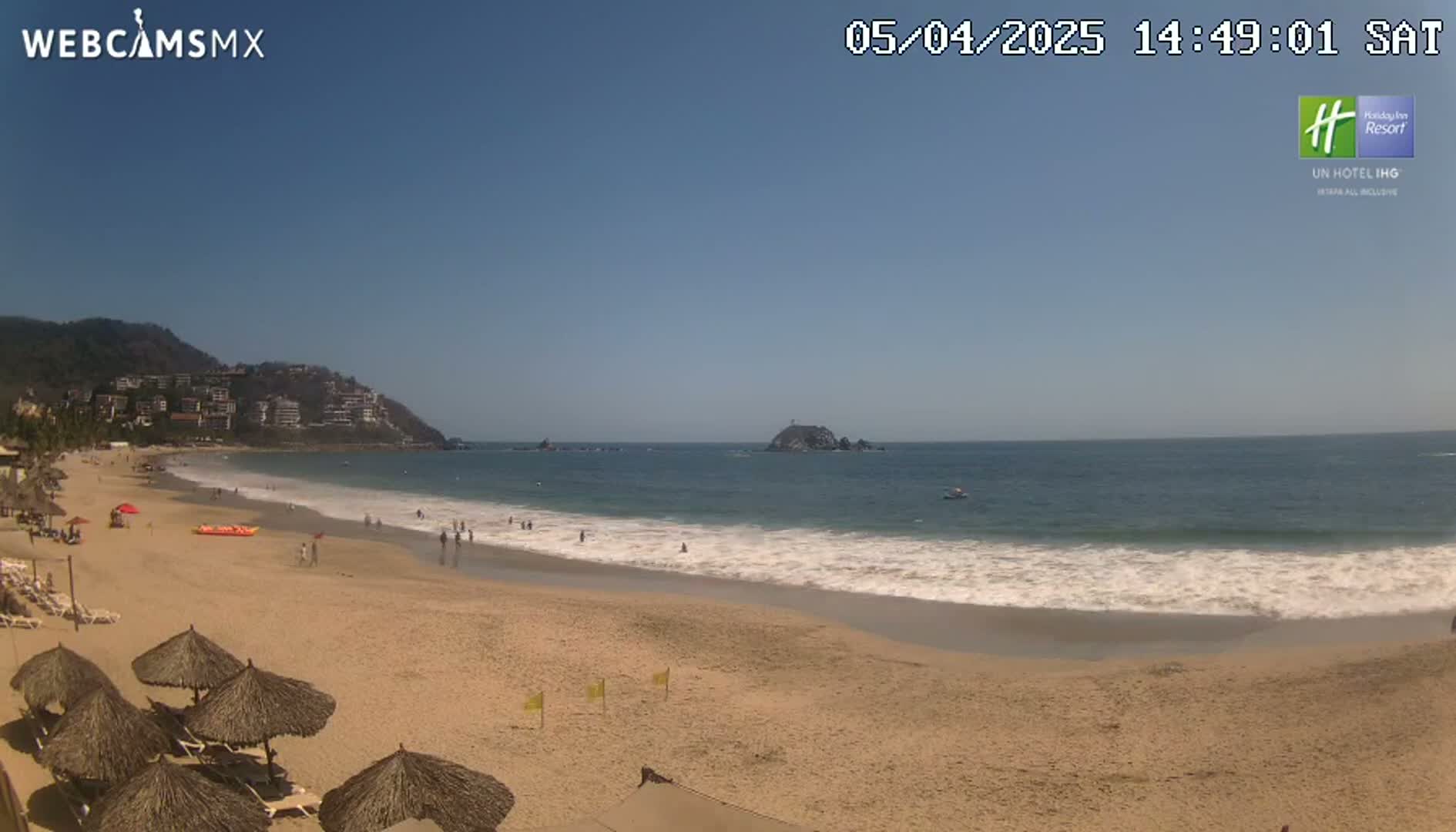A sandy beach with calm waves, a few people swimming and lounging, and thatch-roofed umbrellas under a clear blue sky.