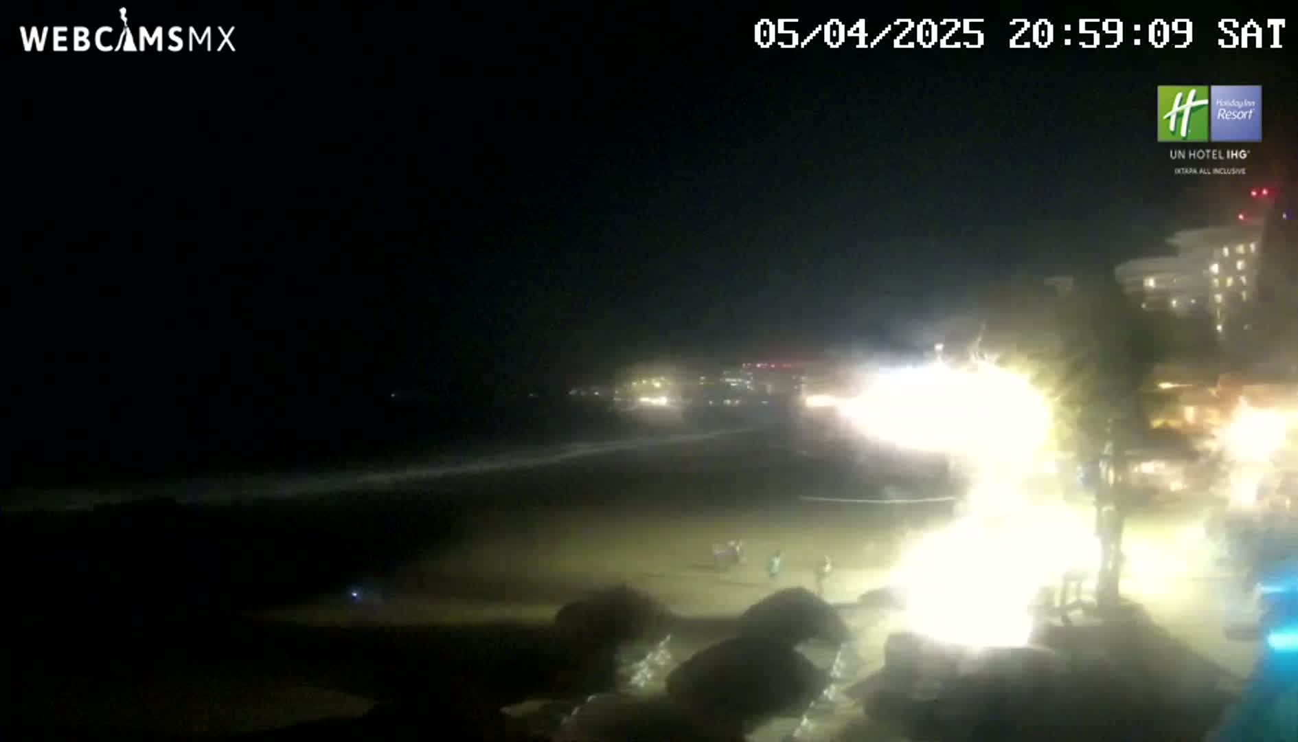 A nighttime beach scene shows a bright light display near the shore, with a calm ocean and a few people visible on the sand under a dark sky.