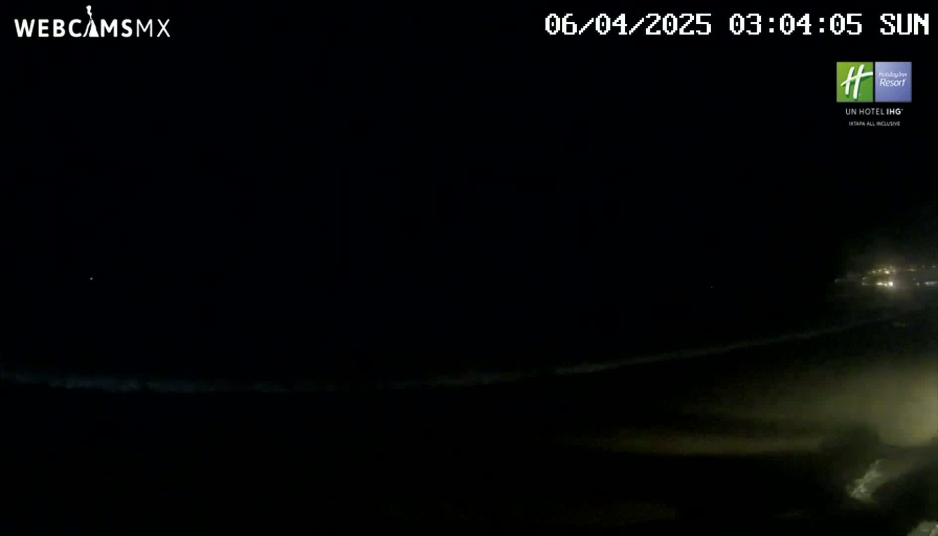 A dark nighttime beach scene shows a dimly lit shoreline and ocean under a clear, dark sky.