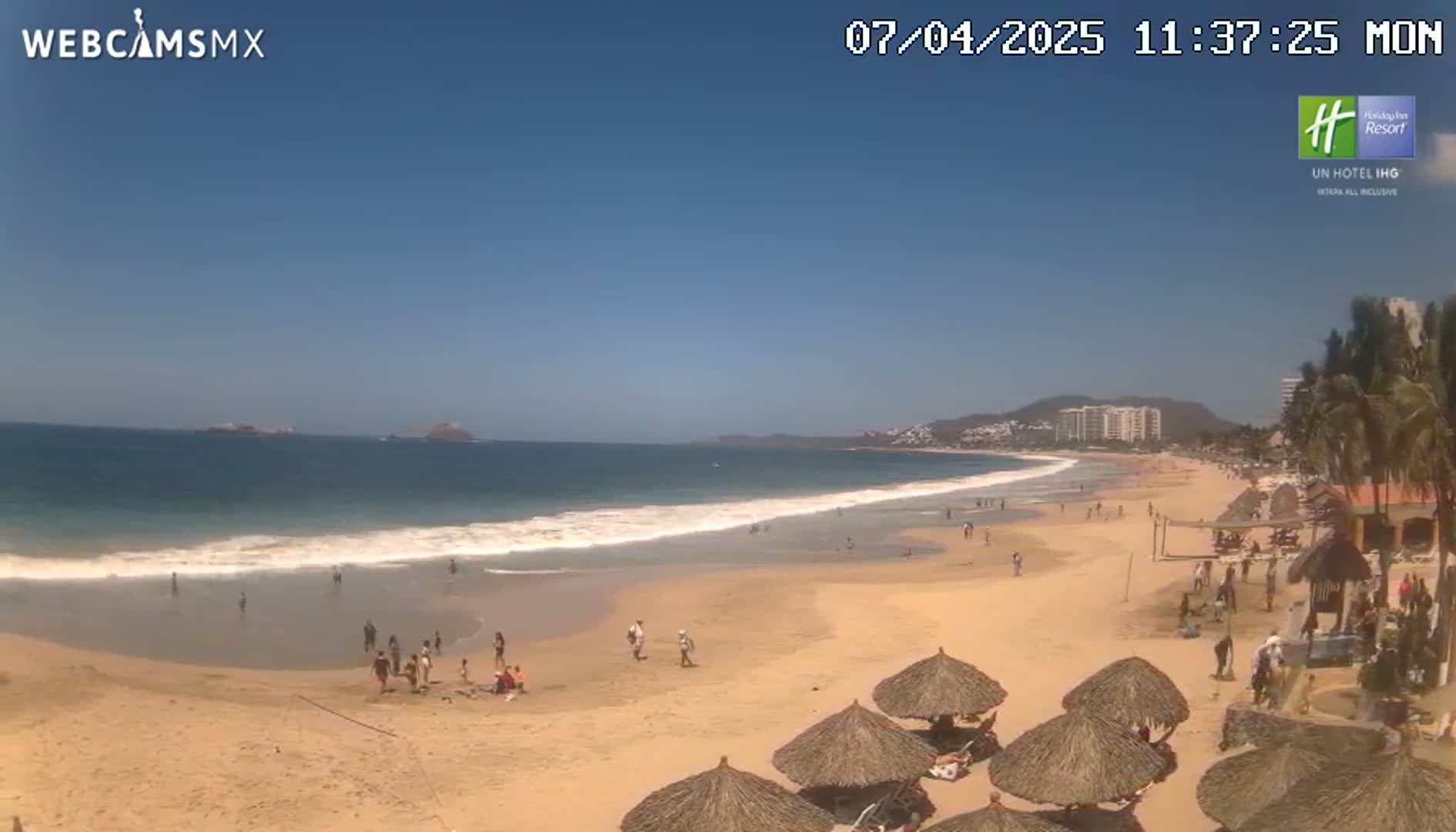A sunny beach with calm waves, scattered beachgoers, and thatch-roofed umbrellas is visible under a clear blue sky.