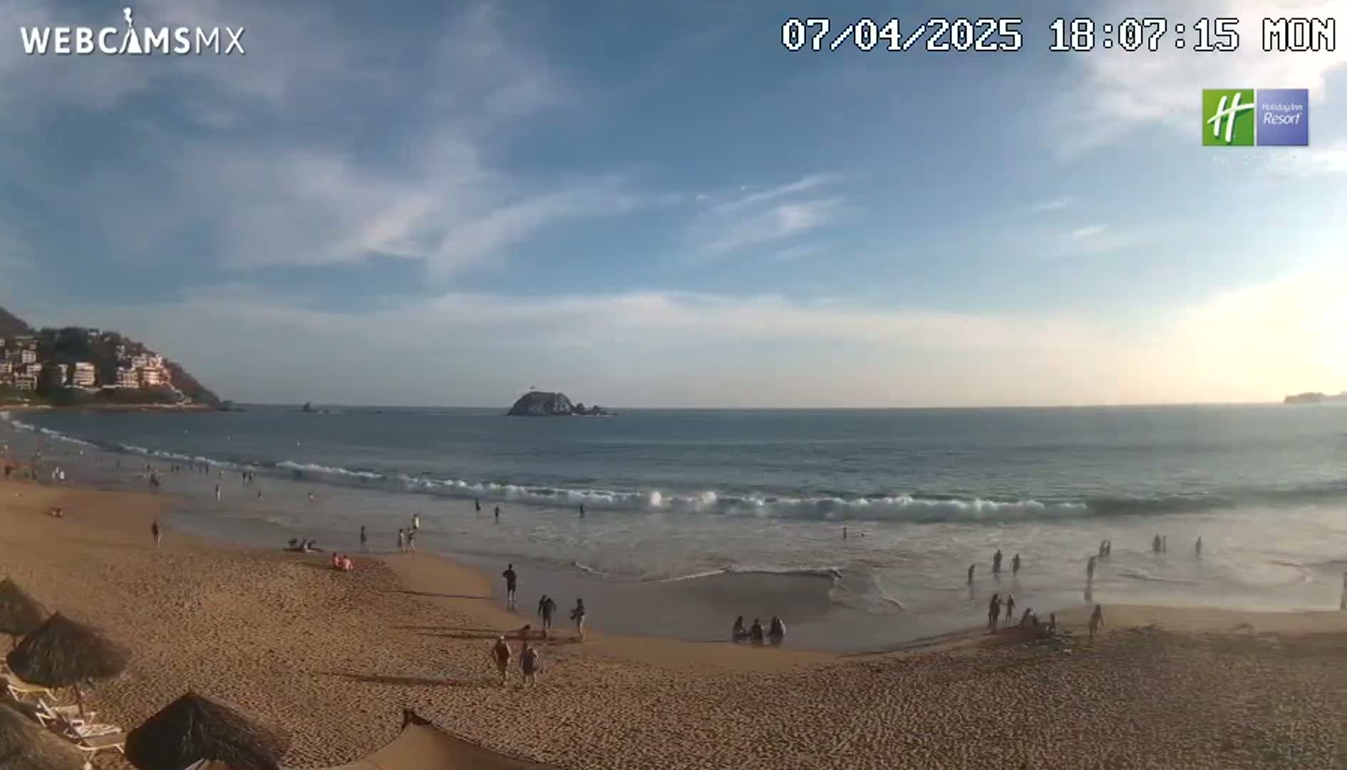 A sandy beach with a few people on it is next to a calm ocean under a partly cloudy sky.
