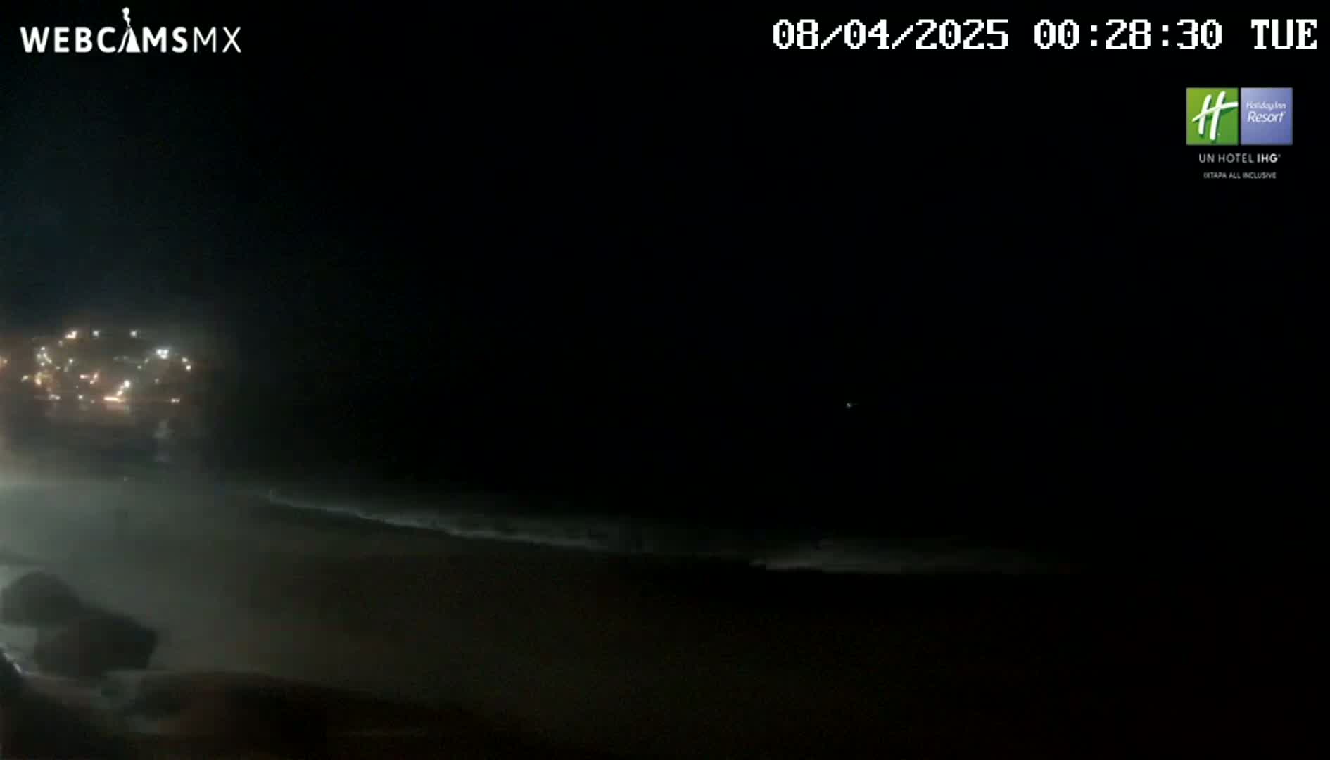 A nighttime view shows a hazy beach with dimly lit waves and distant city lights under a dark sky.