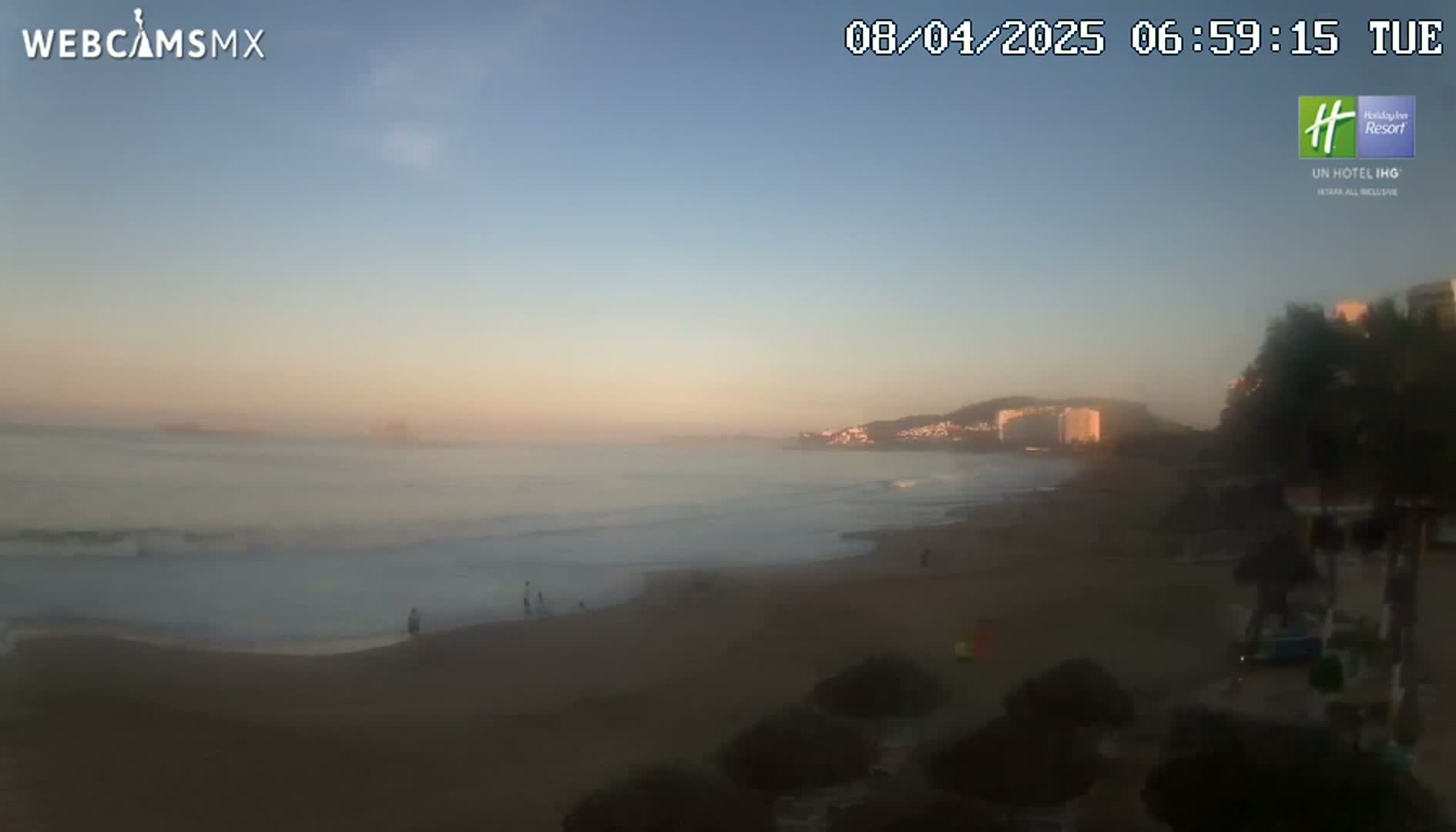 A hazy sunrise illuminates a sandy beach with a calm ocean and distant buildings under a clear sky.