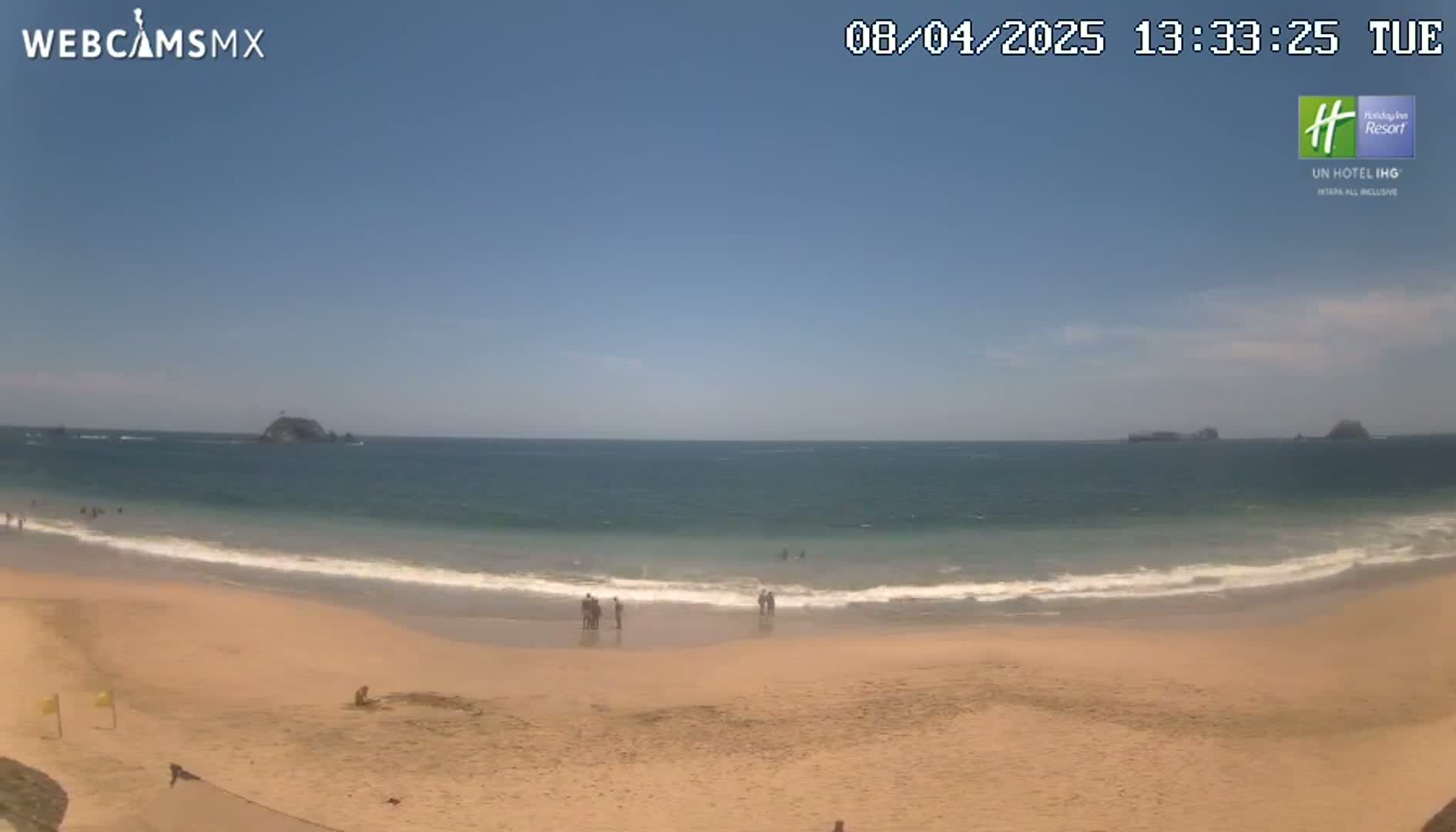 A sandy beach with a few people on it stretches to a calm ocean under a clear blue sky.