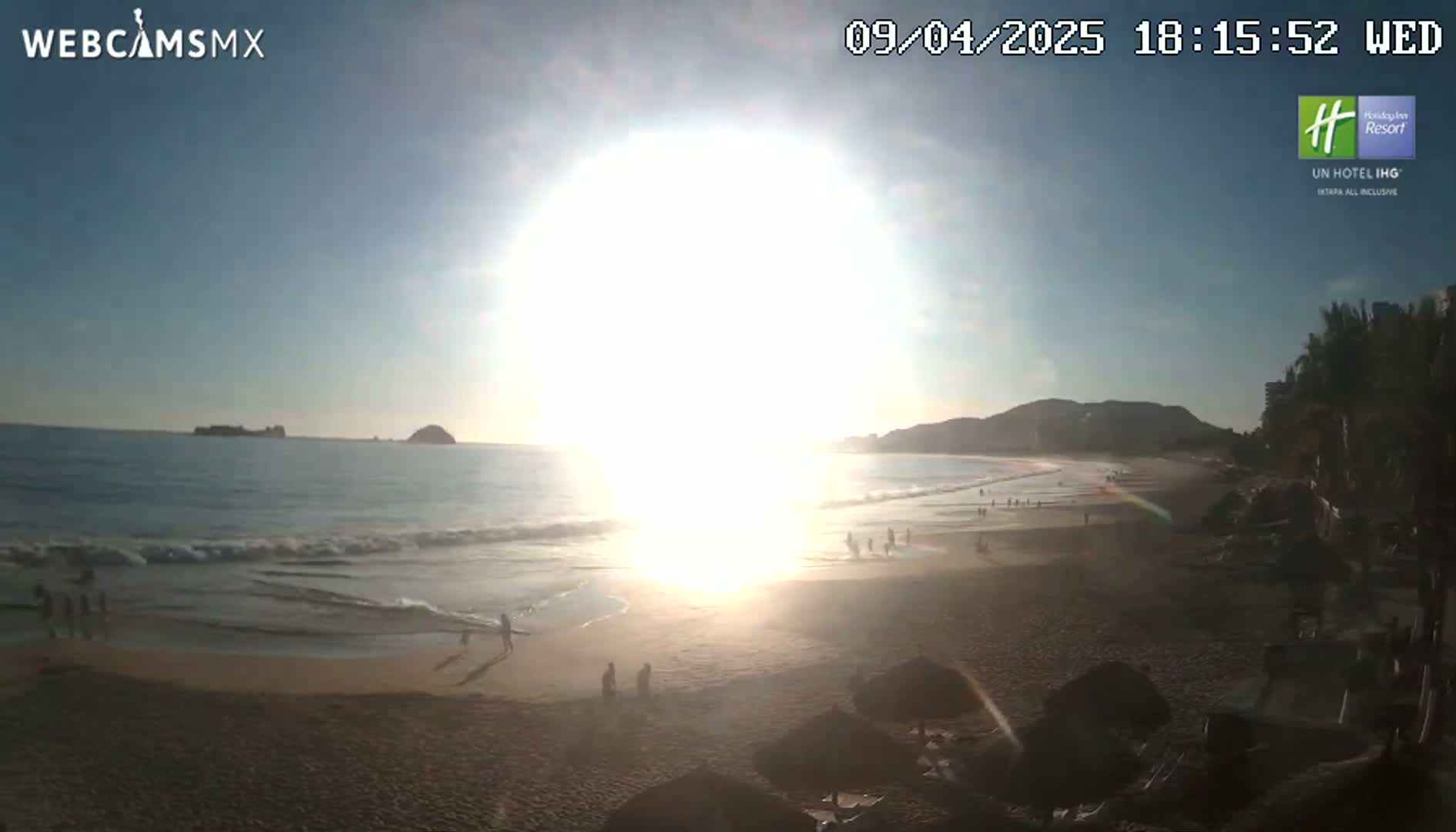 A bright, sunny day at a beach with gentle waves, showing several people walking along the shore.