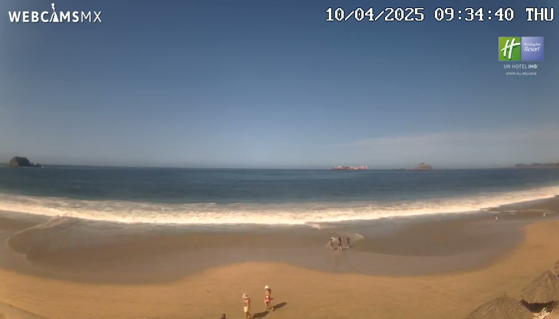 A sandy beach with a few people on it, gentle waves lapping the shore, and a clear blue sky overhead.
