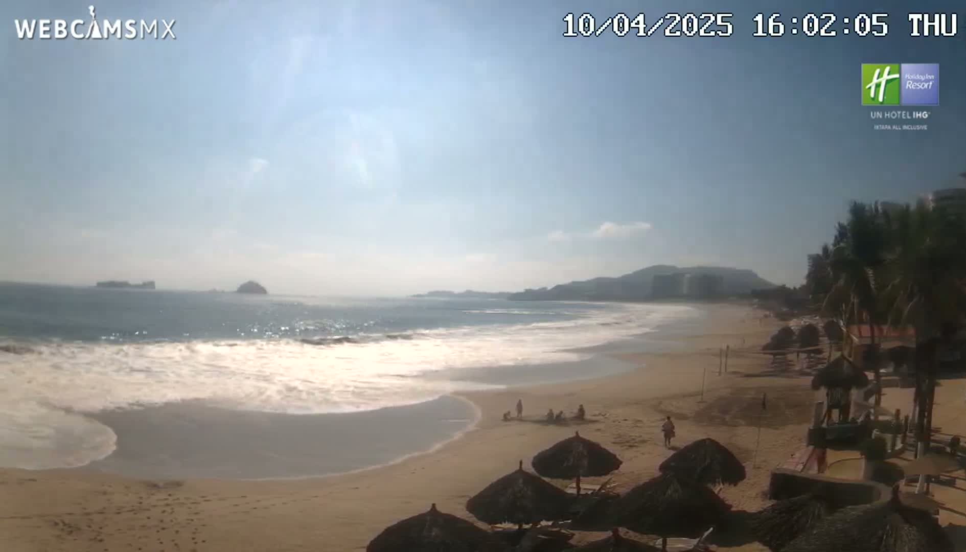 A sandy beach with gentle waves under a mostly clear sky features several beach umbrellas and a few people scattered along the shoreline.